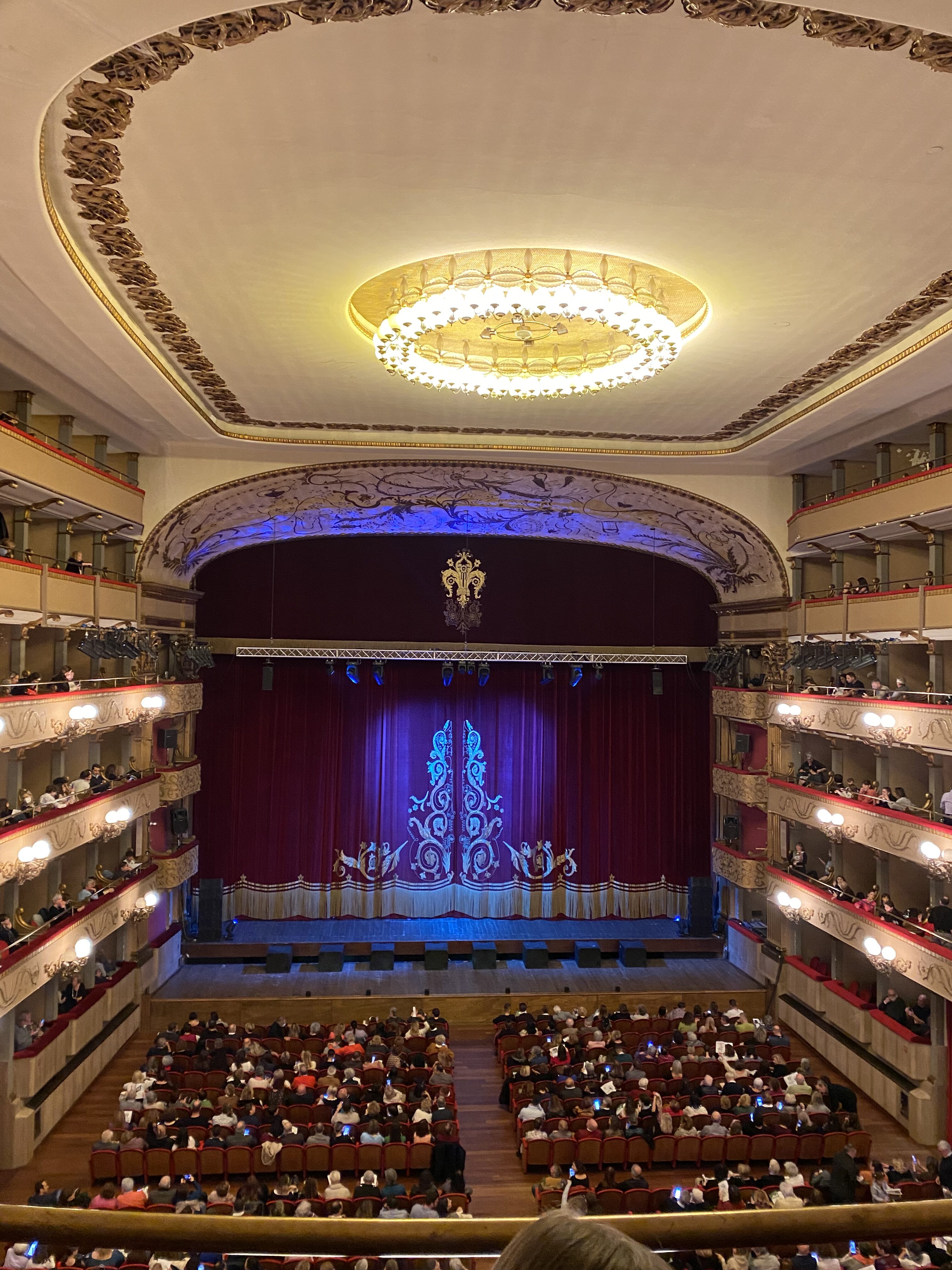 The illuminated interior of Teatro Verdi in Florence with a bright gold chandelier and red curtains covering the stage.