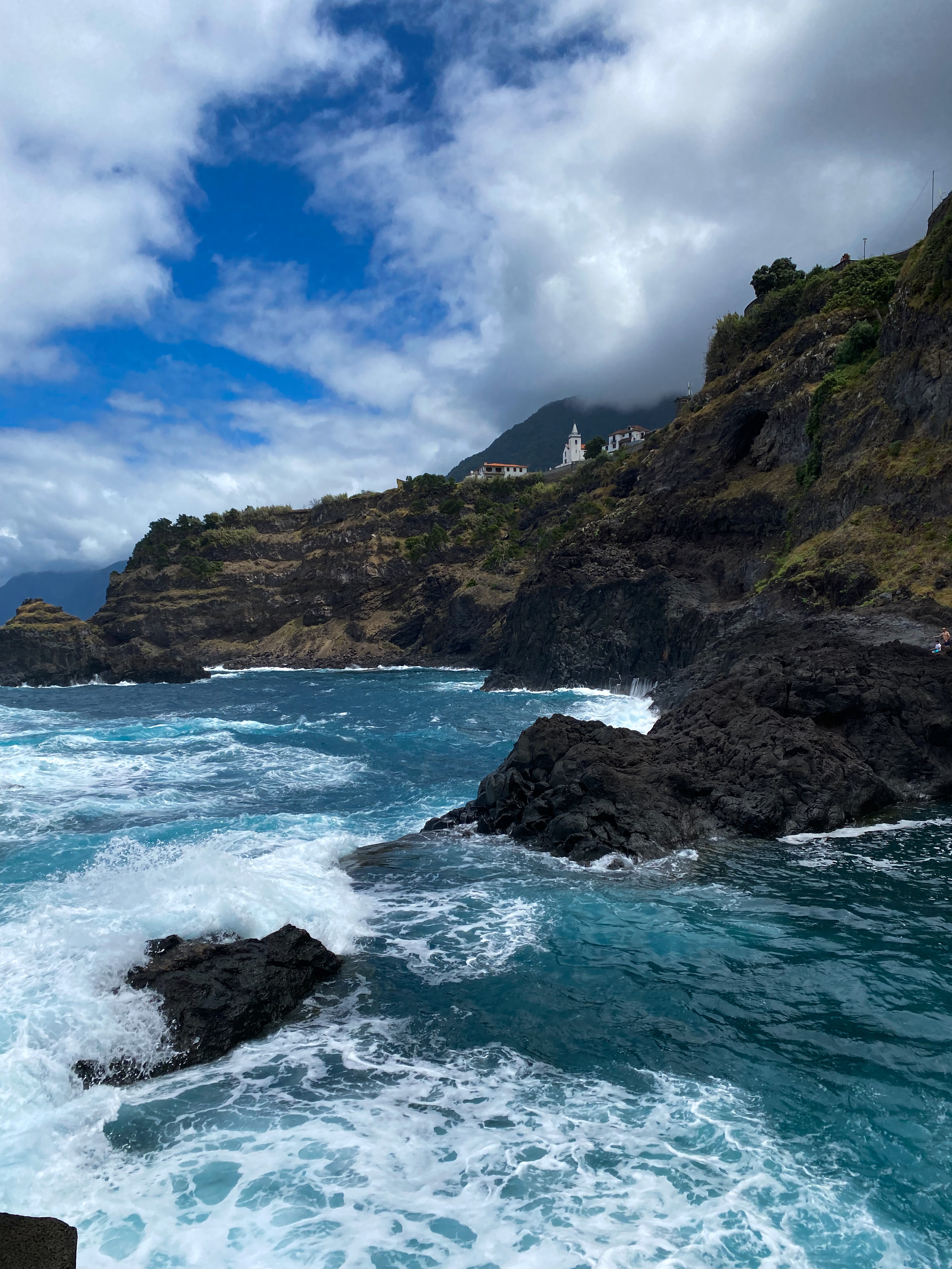 A black-rock cliffside with turquoise blue waters and a cloudy blue sky in Madeira.