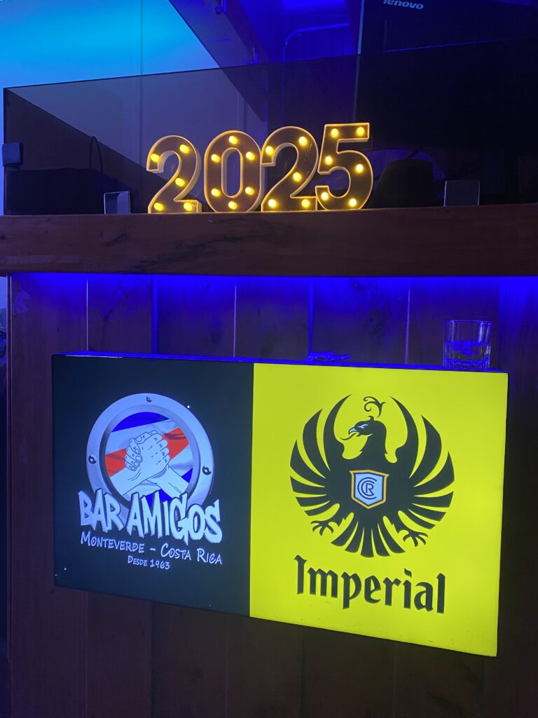 A light sign in a dark bar with the numbers, 2025, above an illuminated sign with the Bar Amigos logo and the Costa Rican beer (Imperial) logo.