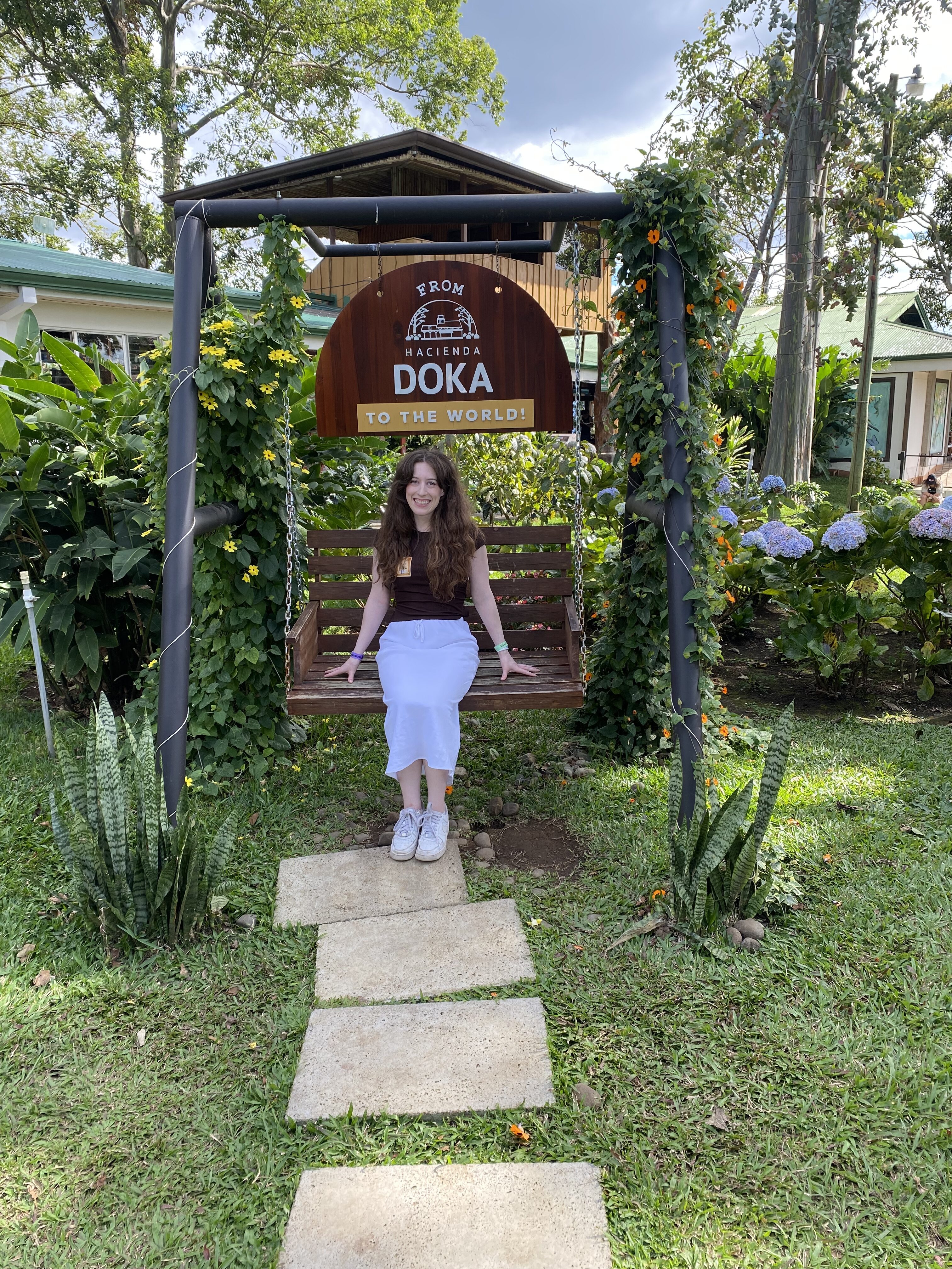 Bella sitting on a swing in a green garden with a sign that says "From Hacienda Doka to the World".