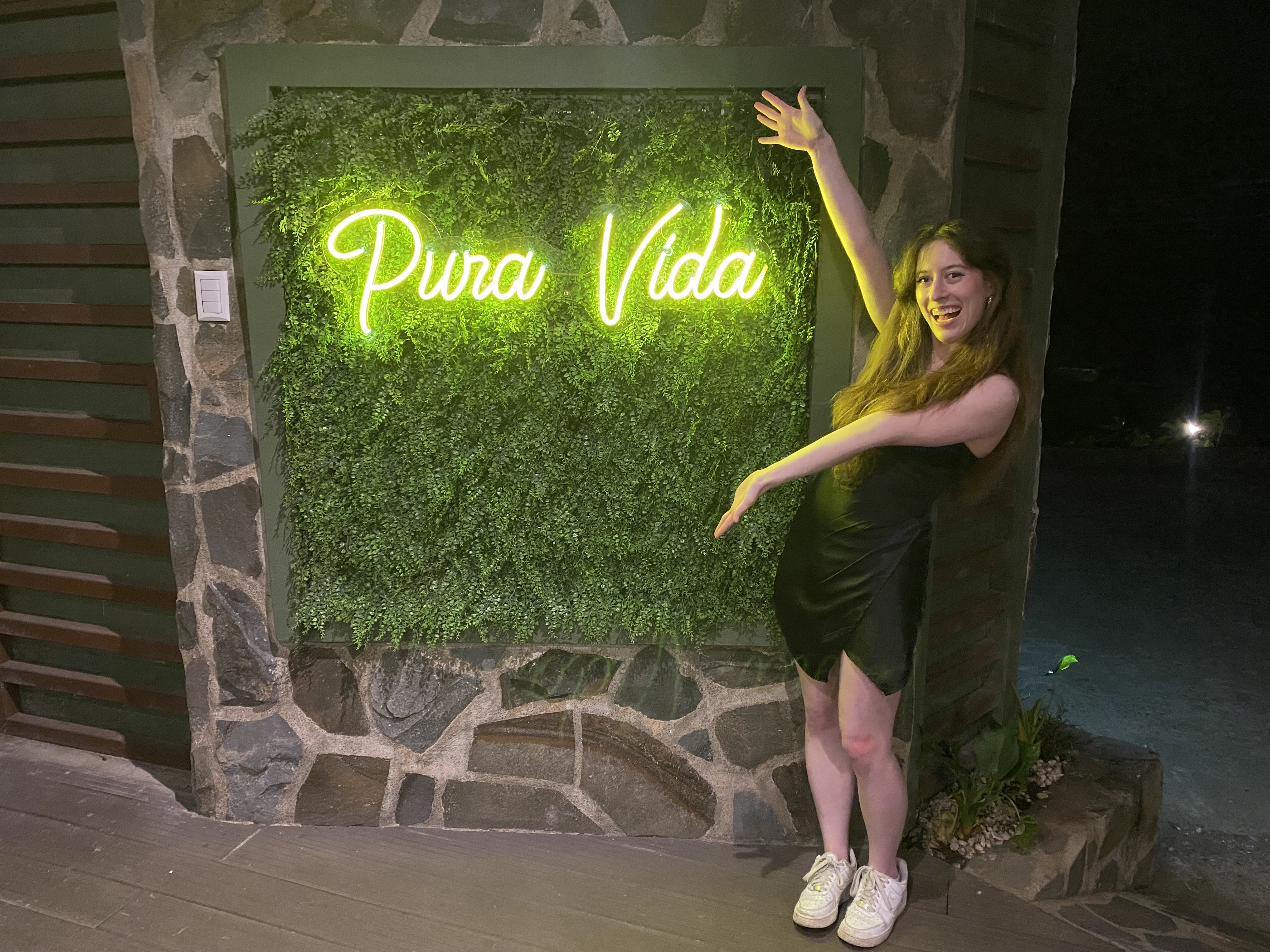 Bella holding her arms out in front of a neon yellow signs that says "Pura Vida".