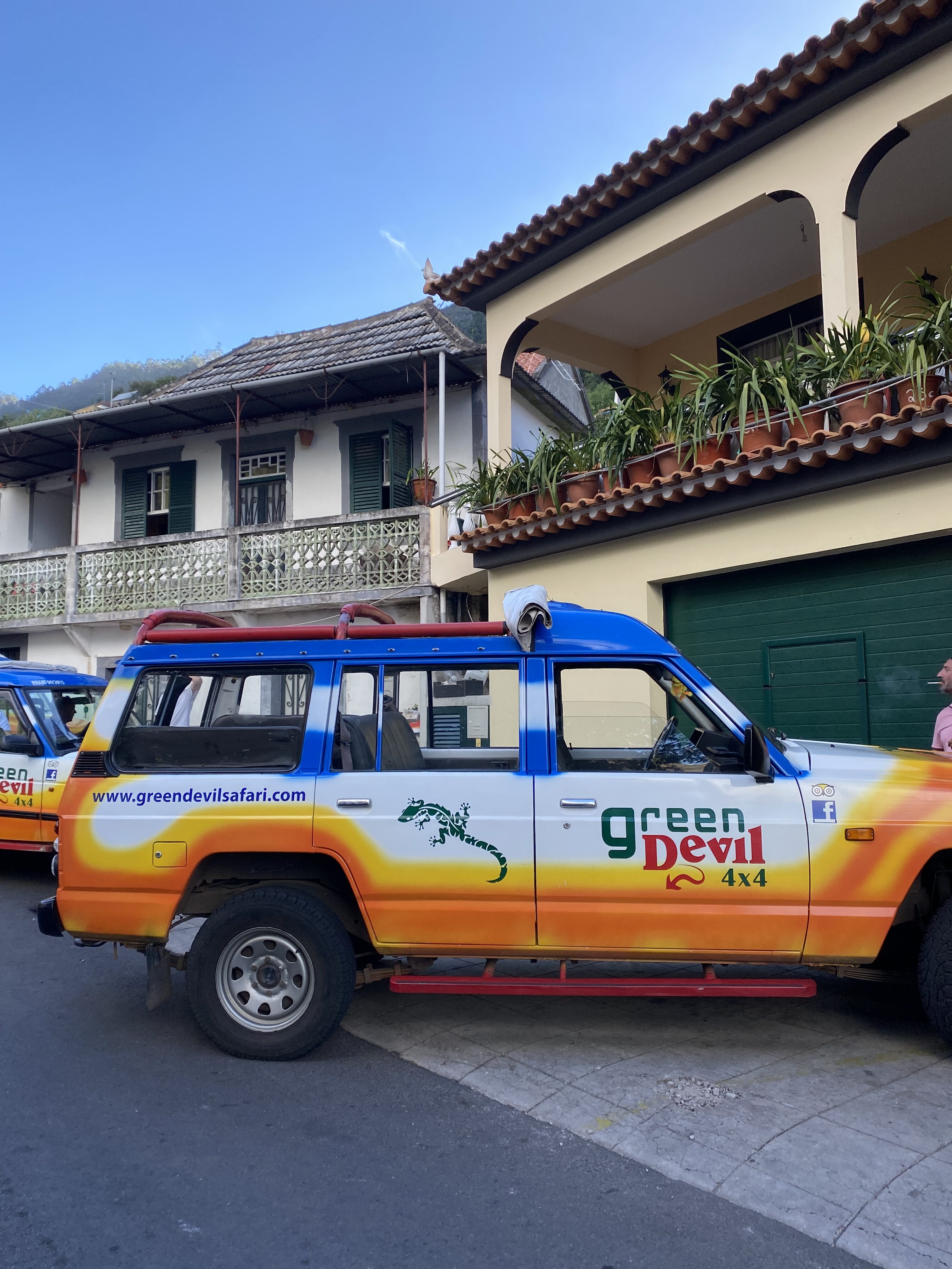 A 4WD vehicle painted with orange, white a blue, labeled with "Green Devil Safari" – the name of the tour operator. The car is parked in front of a villa with a tan balcony with flowerpots.