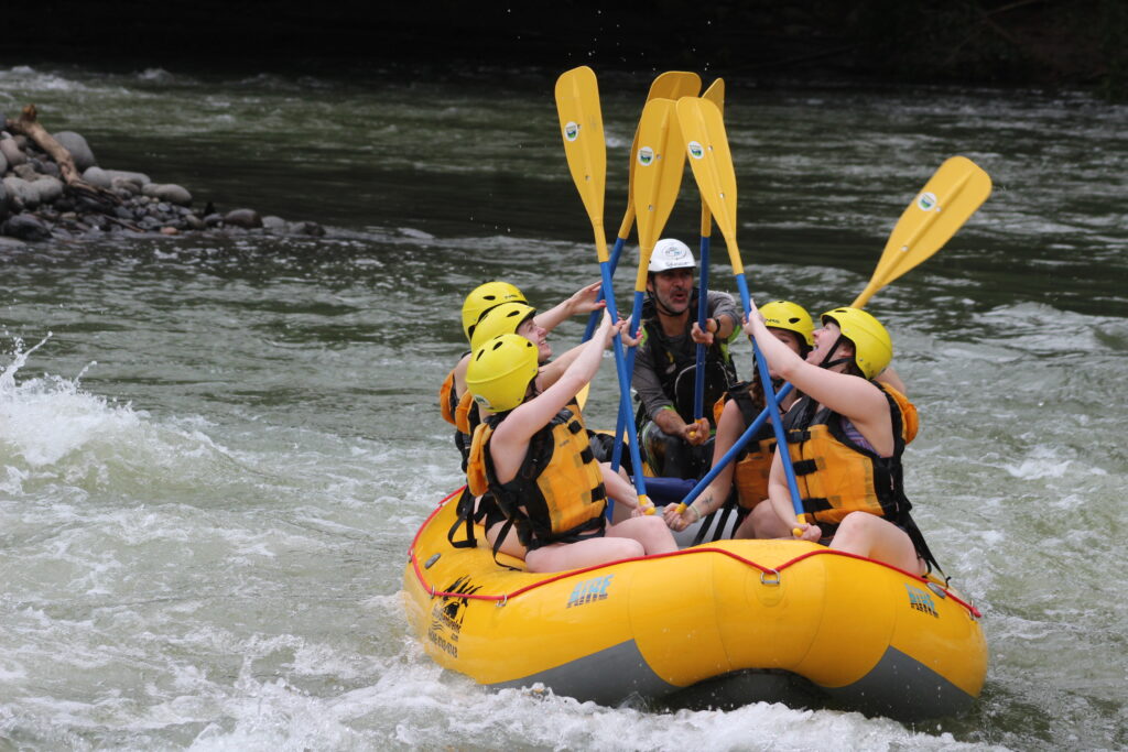 A group of young travelers whitewater rafting, holding their paddles up to the sky.