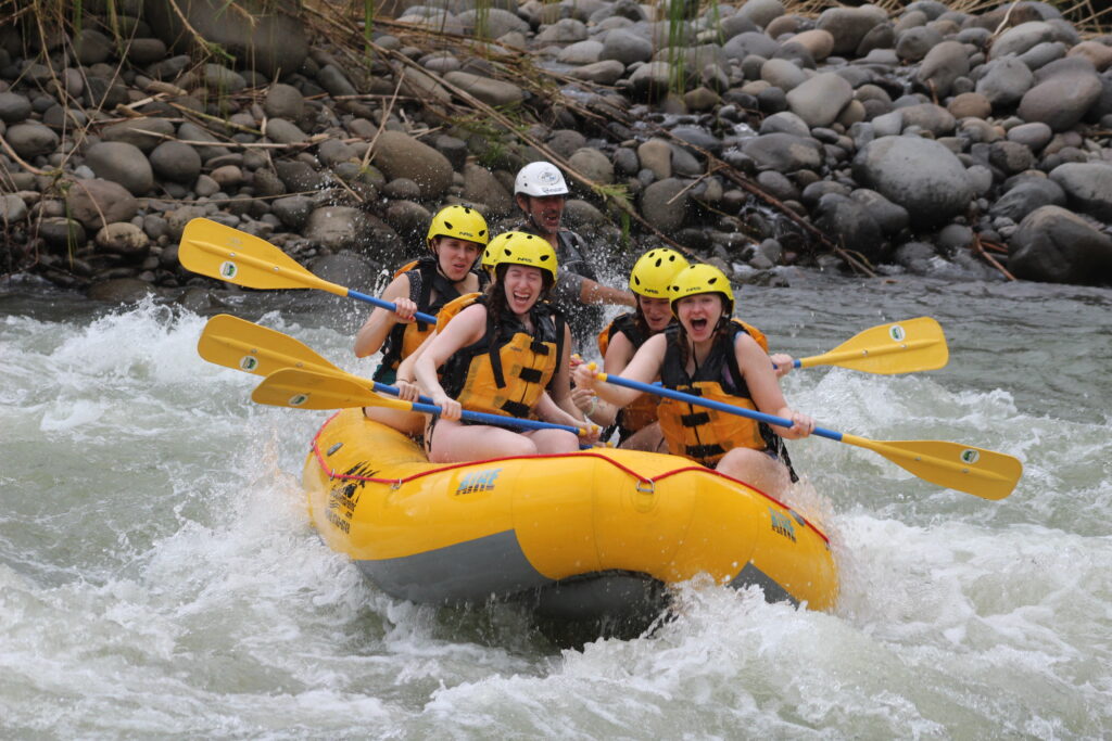 A yellow rafting splashing in a raging river with a group of travelers holding yellow paddles. The group is shocked with the splash of the river.