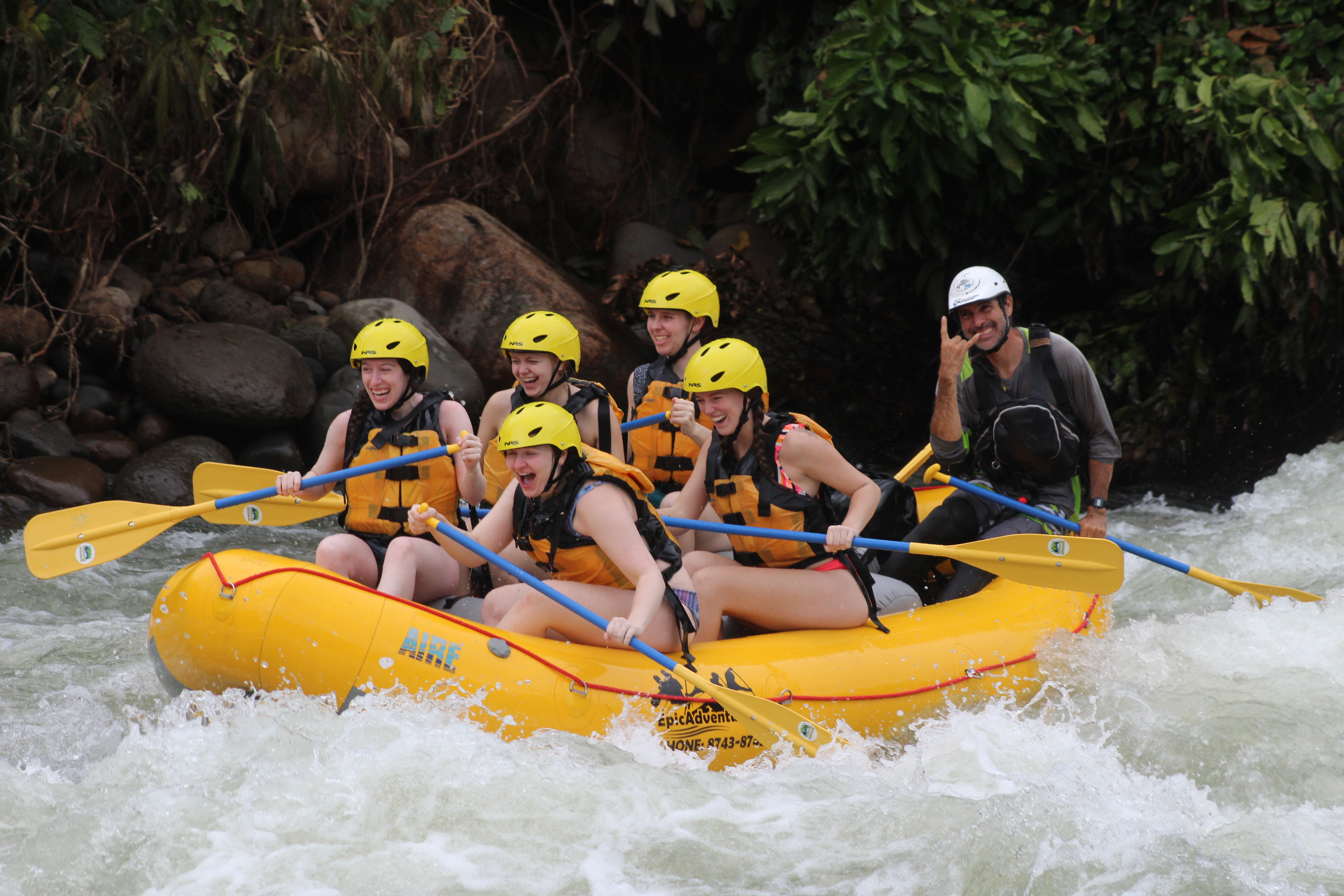 A group of young adventurers on a yellow raft, cruising along the rapids of the Sarapiqui River. Their guide is holding up a "rock on" hand signal at the back of the raft.