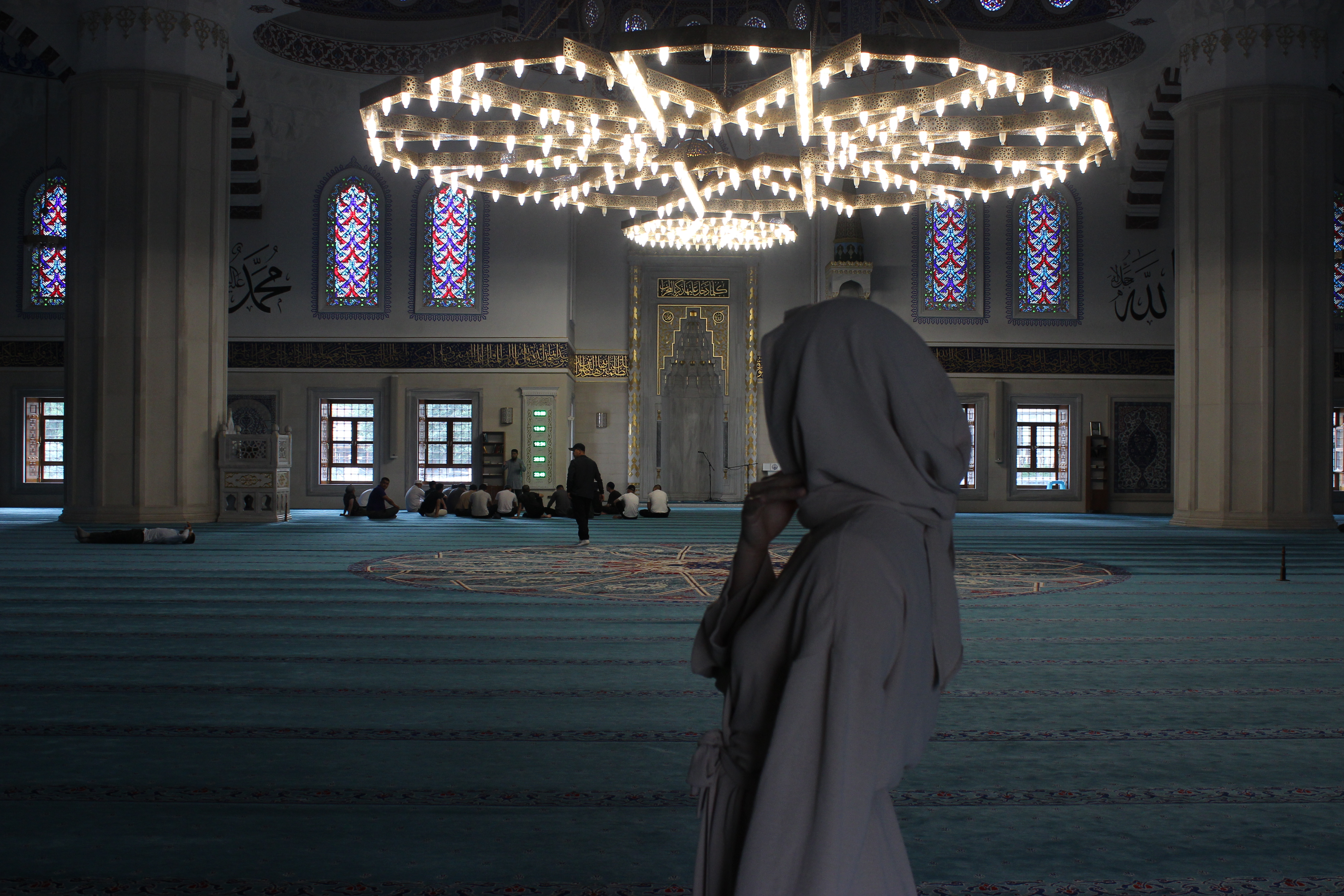 A woman wearing a full head covering in Bishkek Central Mosque, gazing upon the darkened prayer room with the bright chandelier.