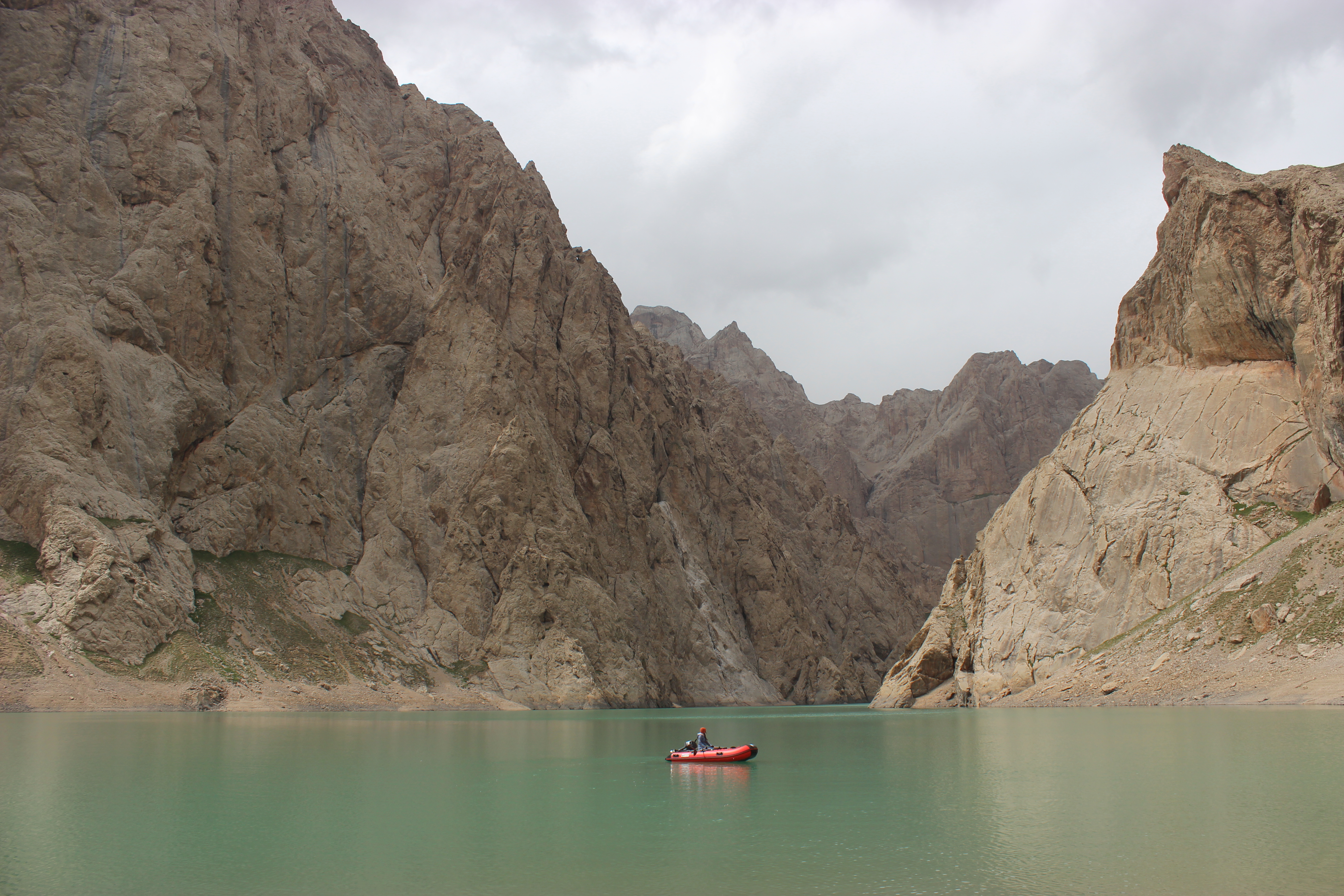 A small motor boat in the center of a greenish/turqoise lake, surrounded by grey rock formations.