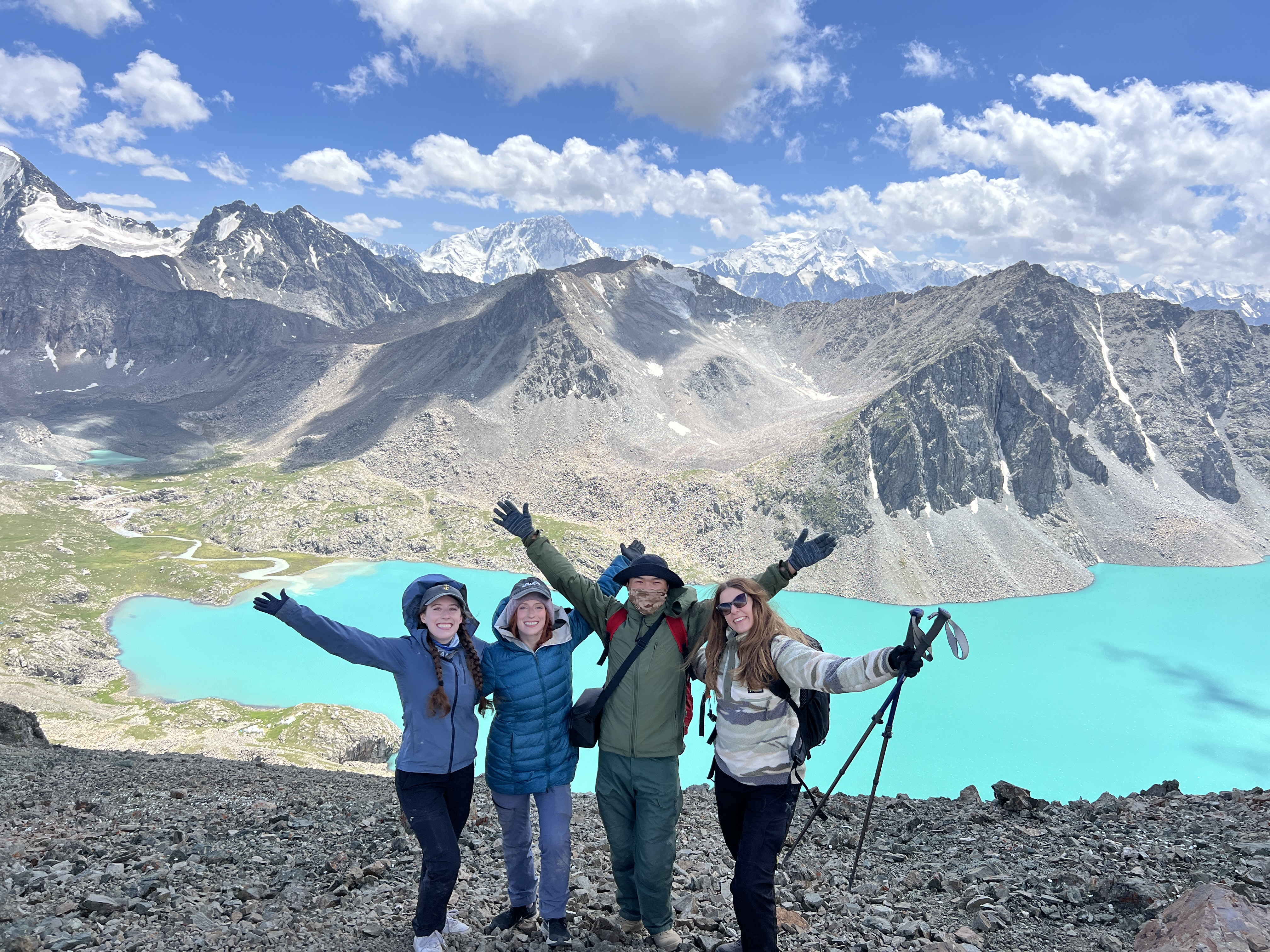 Bella, Lily, Medet and Lena, with their arms held out wide at the top of Ala Kul pass. The vibrant lake shines below.