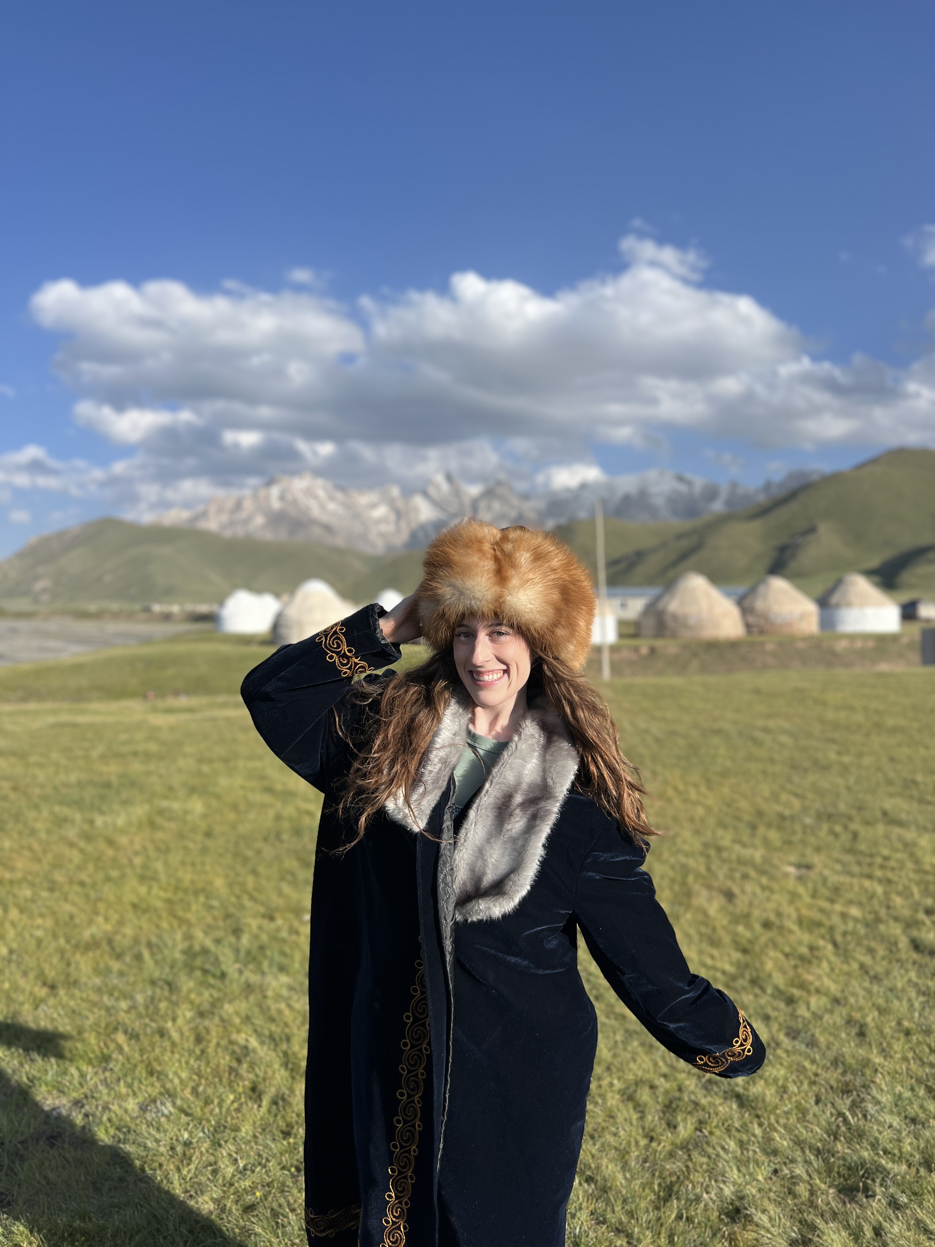 Bella, posing in a long, blue coat and a brown fur hat, traditional Kyrgyz clothing. She is standing in front of a yurt camp in the Kok Kiya valley.