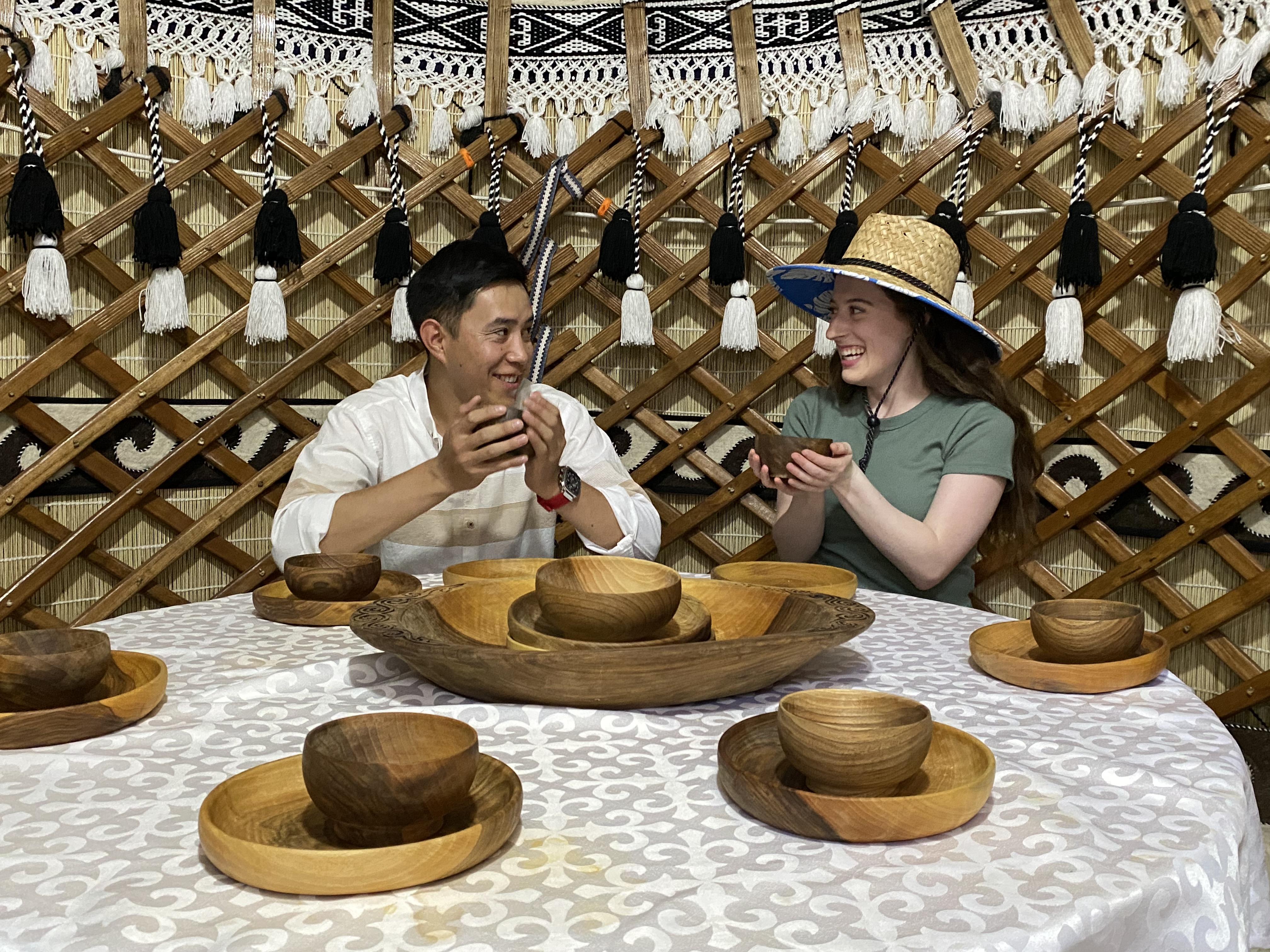 A young man and young women wearing a big straw hat sitting at a round table in a yurt, holding wooden bowls.