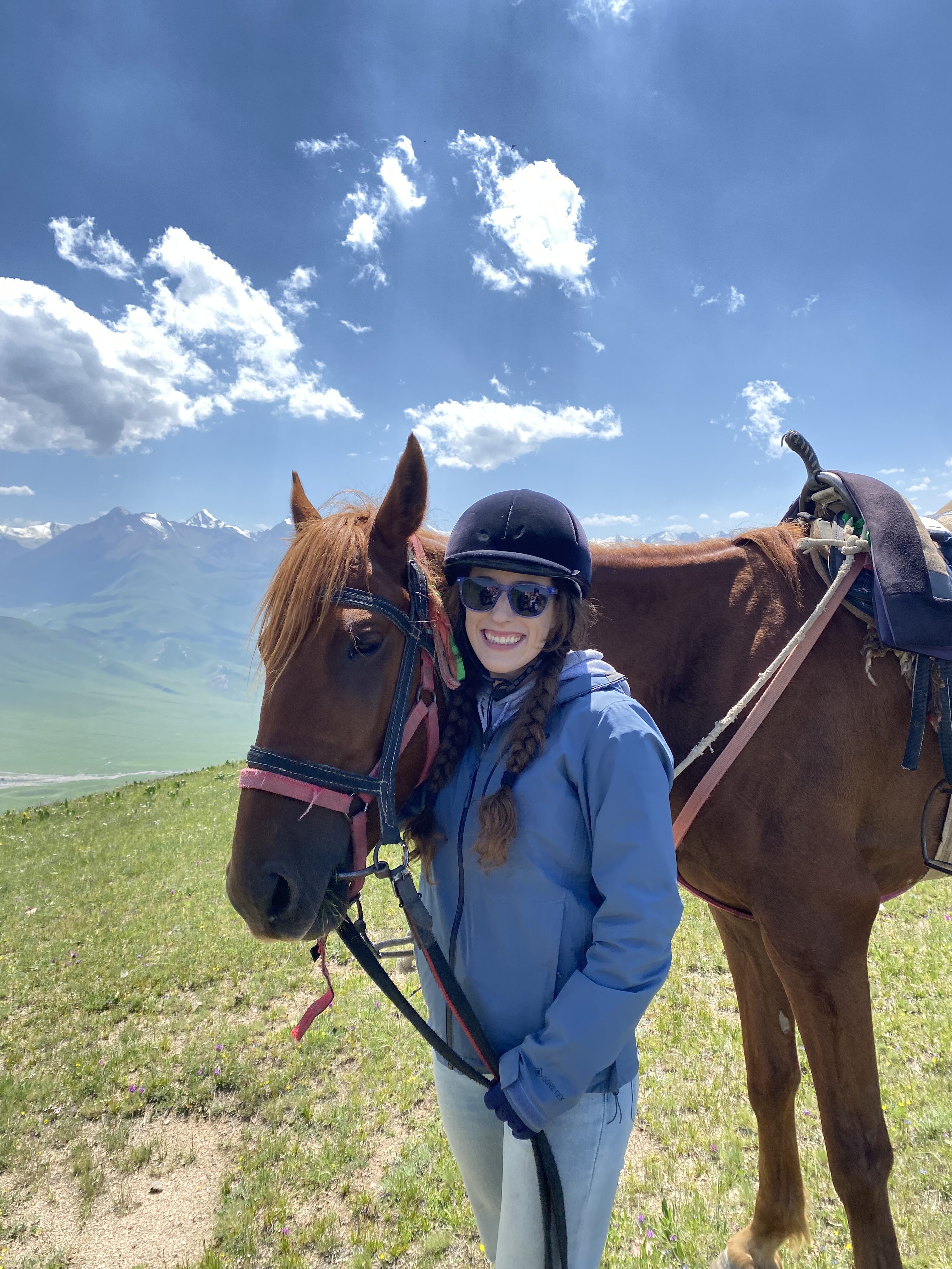 Bella stands beneath the chin of her tall horse named Alamo, at the top of a mountain pass in Kyrgyzstan.
