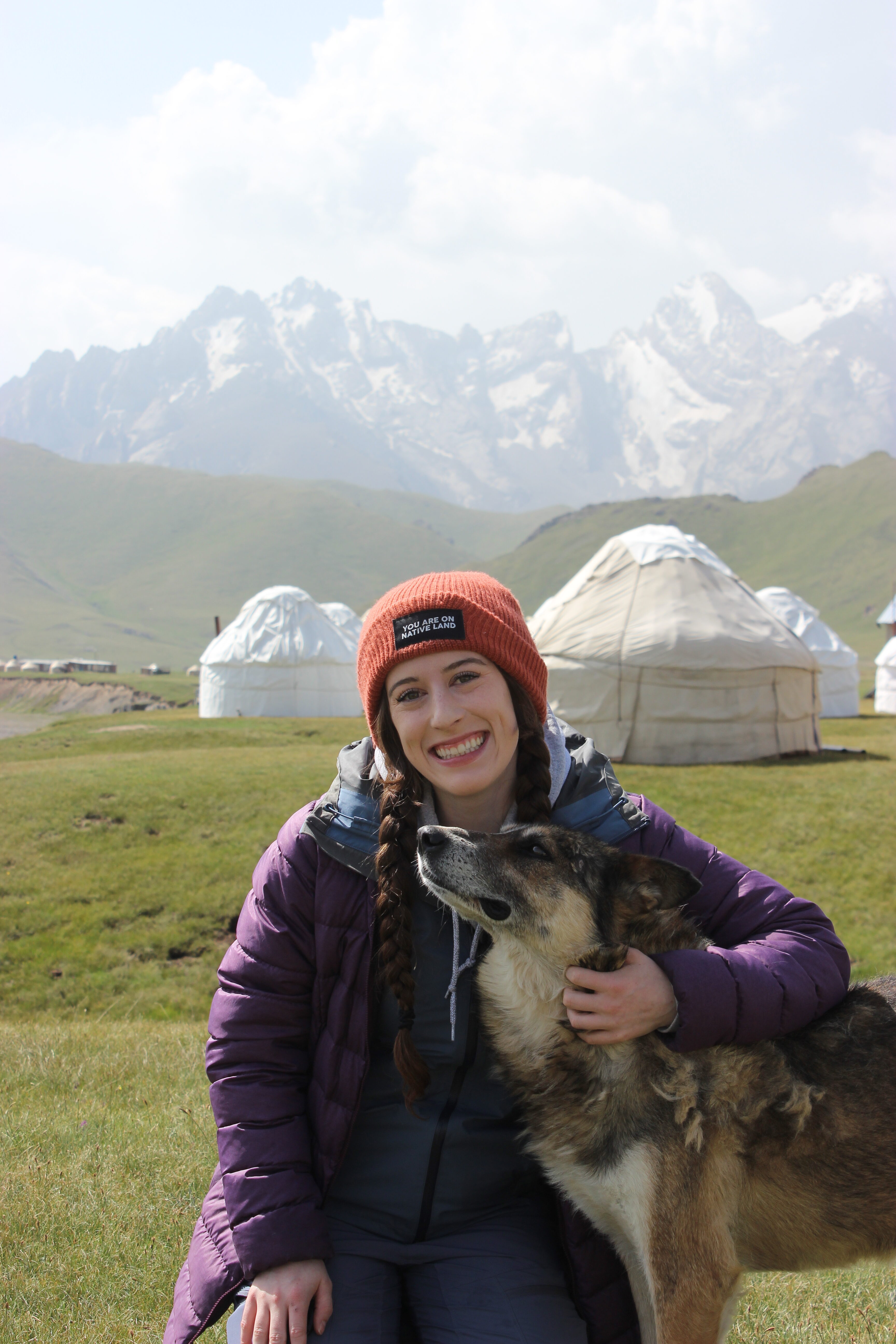 Bella, wearing a purple coat and an orange hat, hugs a dog in front of the yurt camp in Kok Kiya valley.