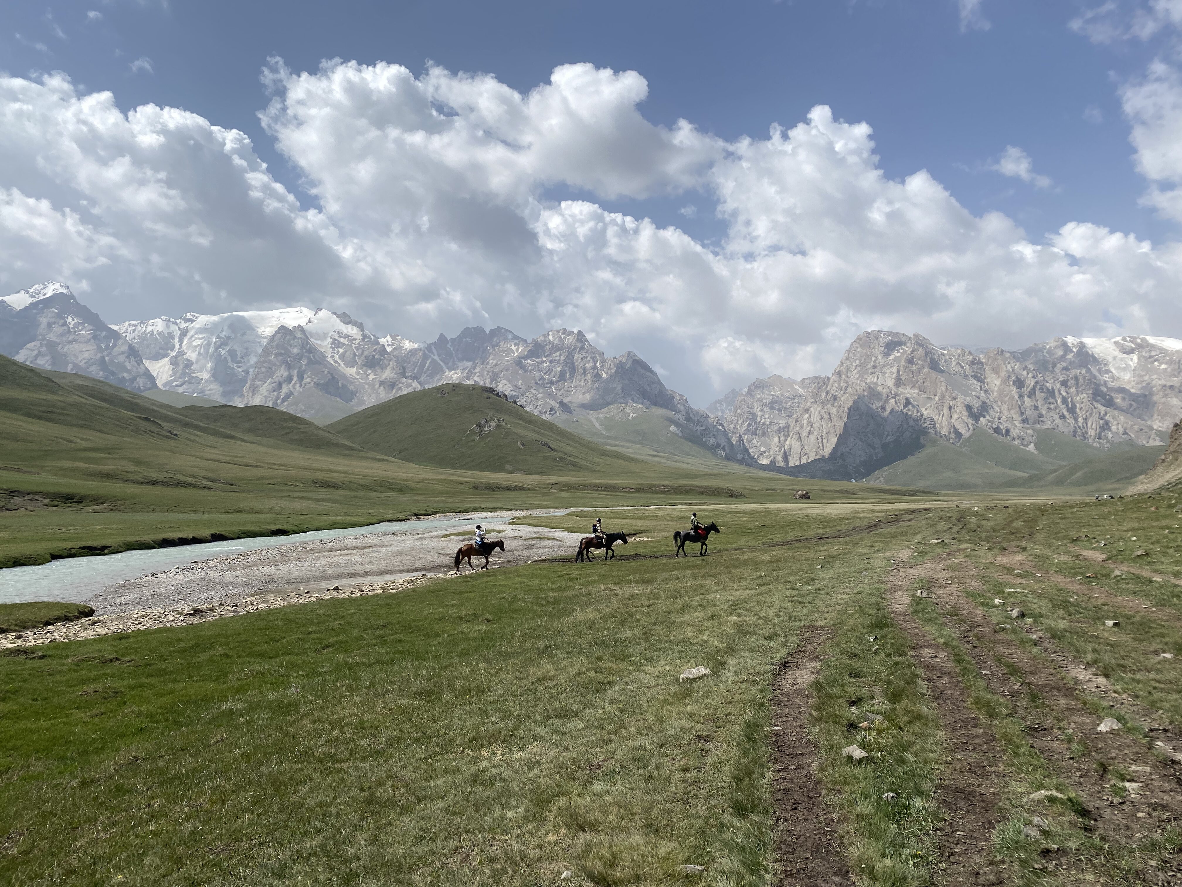 Three horseback riders traversing the grassy plains in Kyrgyzstan, heading towards jagged peaks that surround Kel Suu lake.