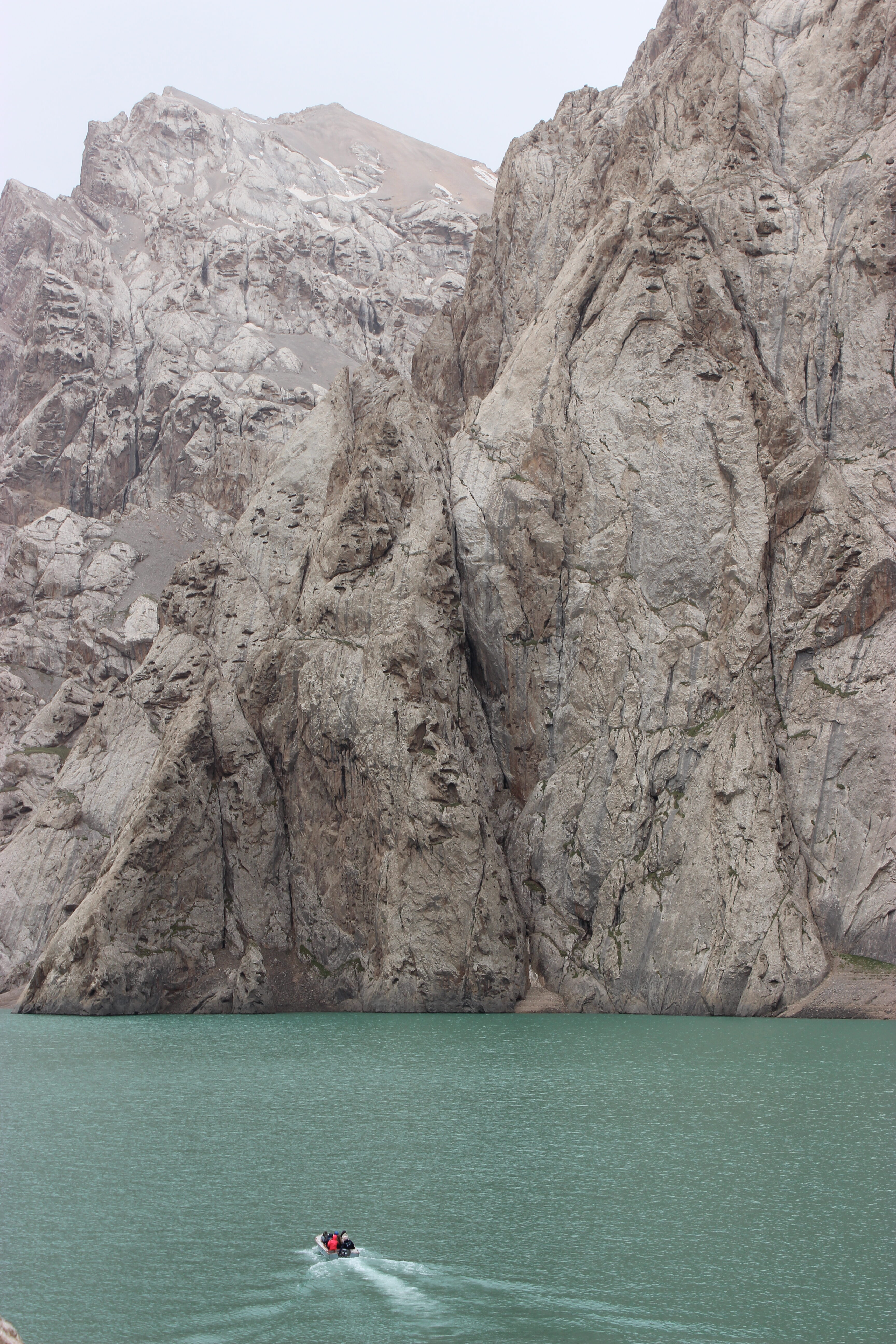 Jagged grey rocks at Kel Suu lake towering above the turquoise water, with a boat cruising along the surface.
