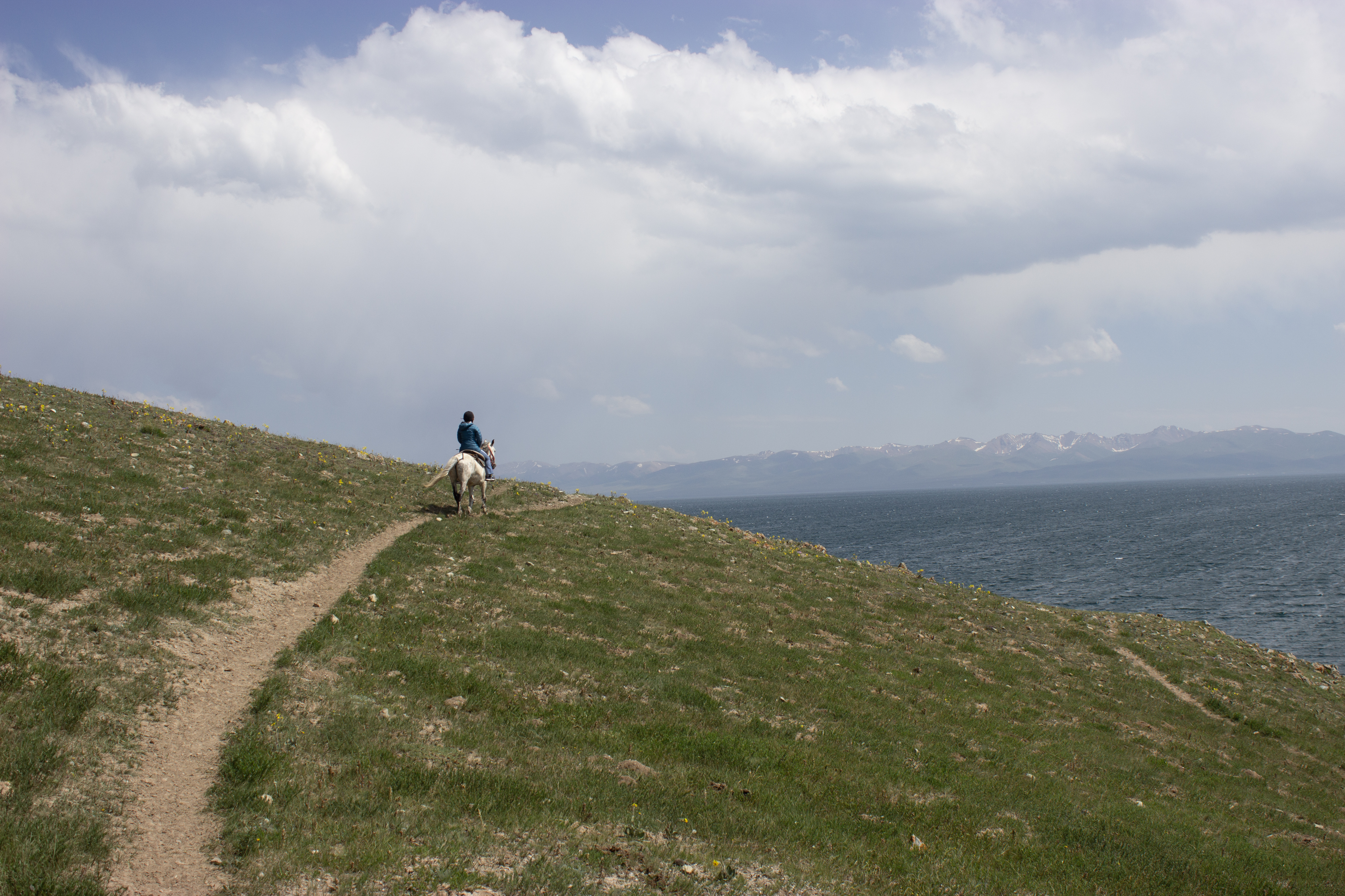 A single horseback rider along a grassy trail, with Song Kul lake in the background.