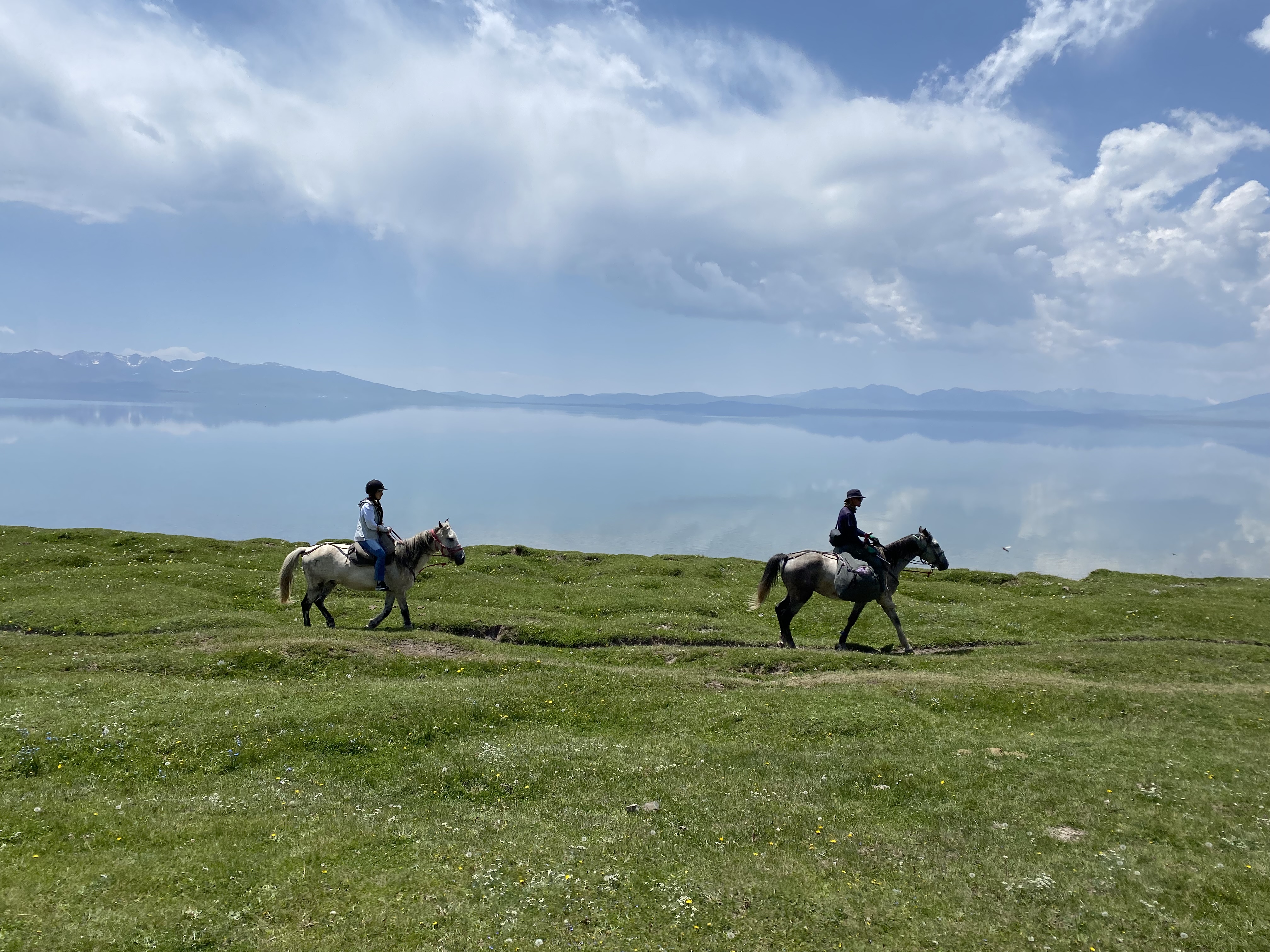 Two horseback ridings, riding along the grassy plains next to Song Kul lake.
