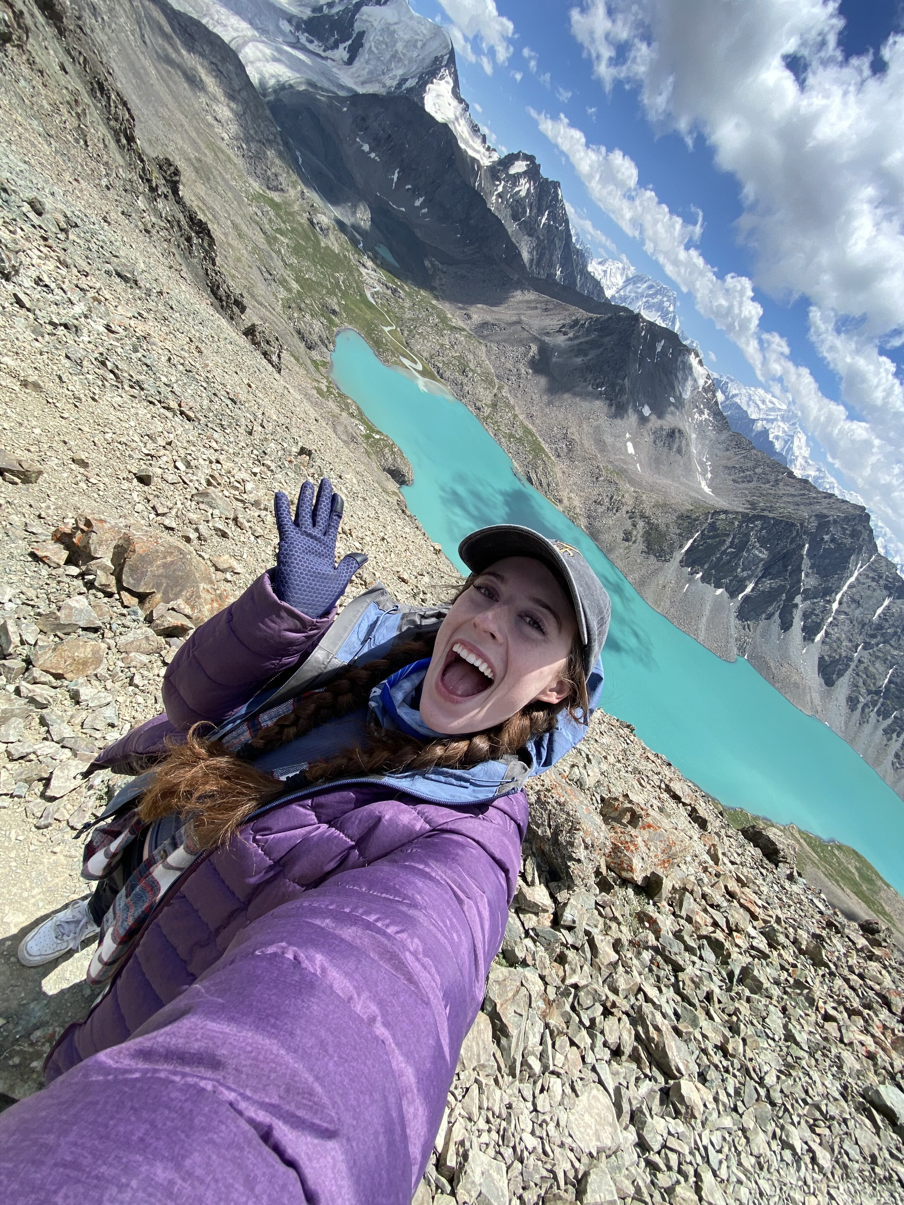 A young woman with two braids and a grey hat has a shocked, open-mouthed expression while holding a gloved-hand out in front of a Rocky Mountain side with an unforgettable blue lake.