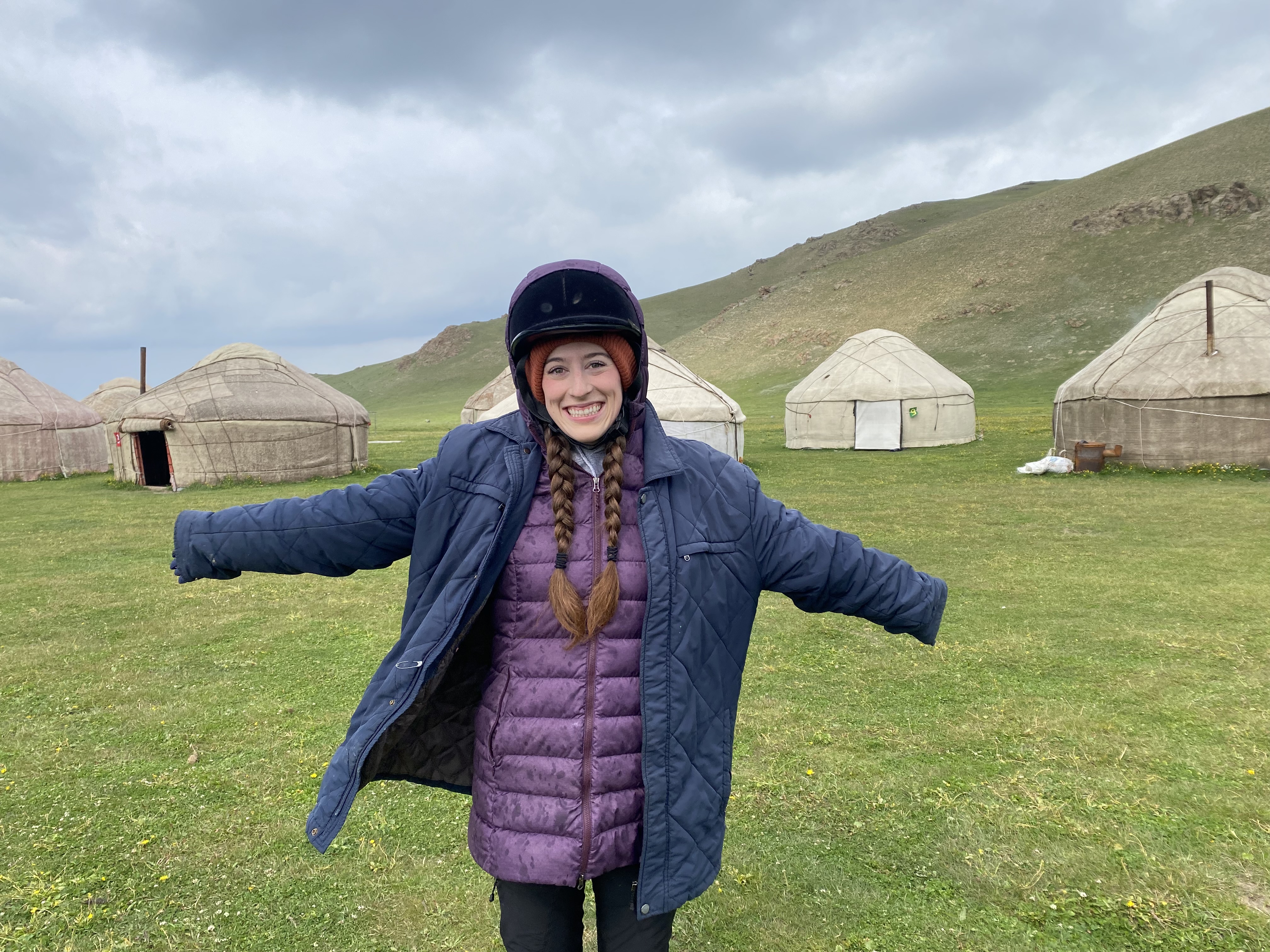 Bella wearing a big blue coat over her purple coat, posing with her arms out in front of a yurt camp near Song Kul.