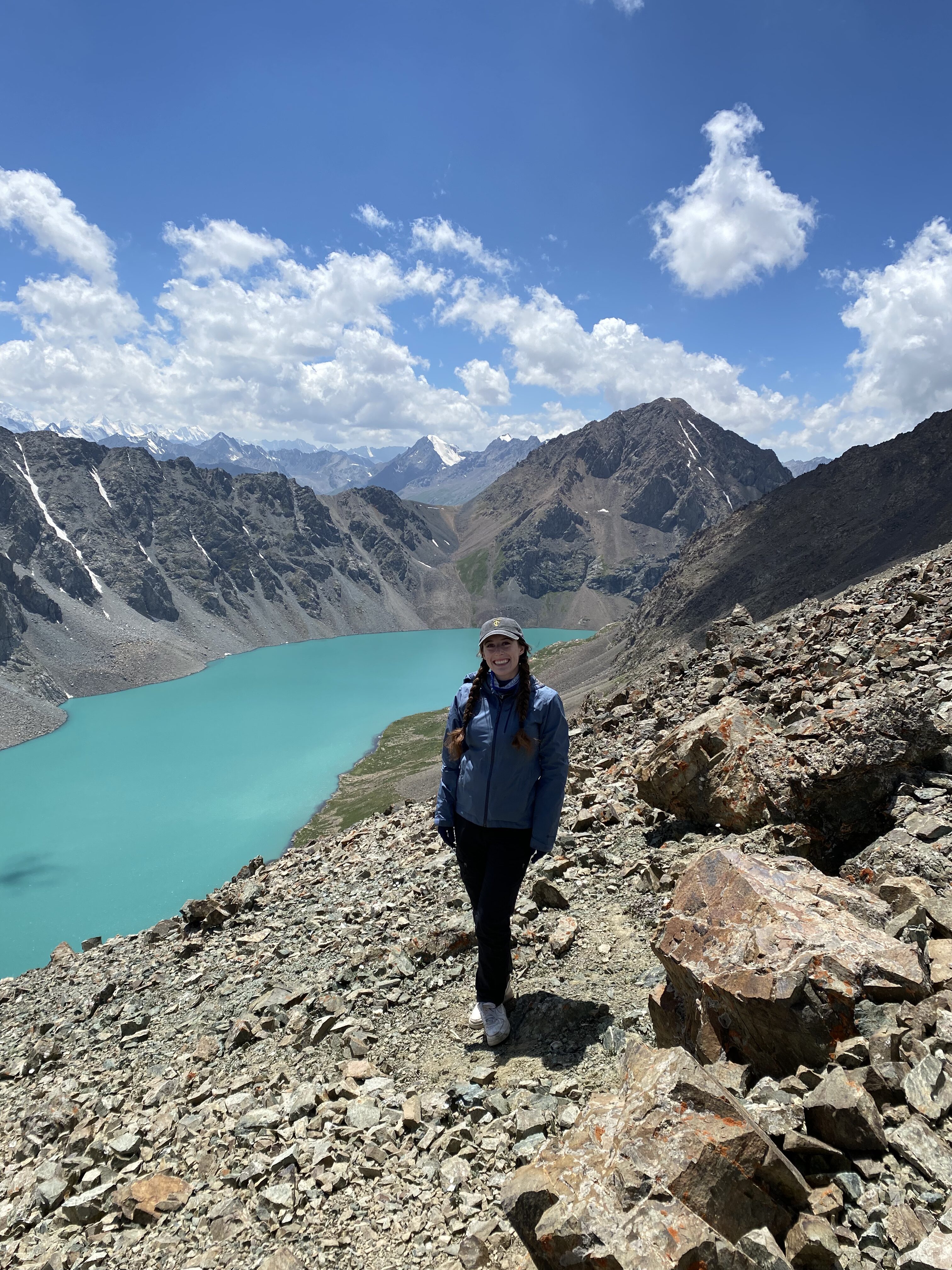 A young woman wearing a blue rain jacket, black pants and a grey ball camp smiles at the top of Ala Kul pass, with the turquoise lake in the background.