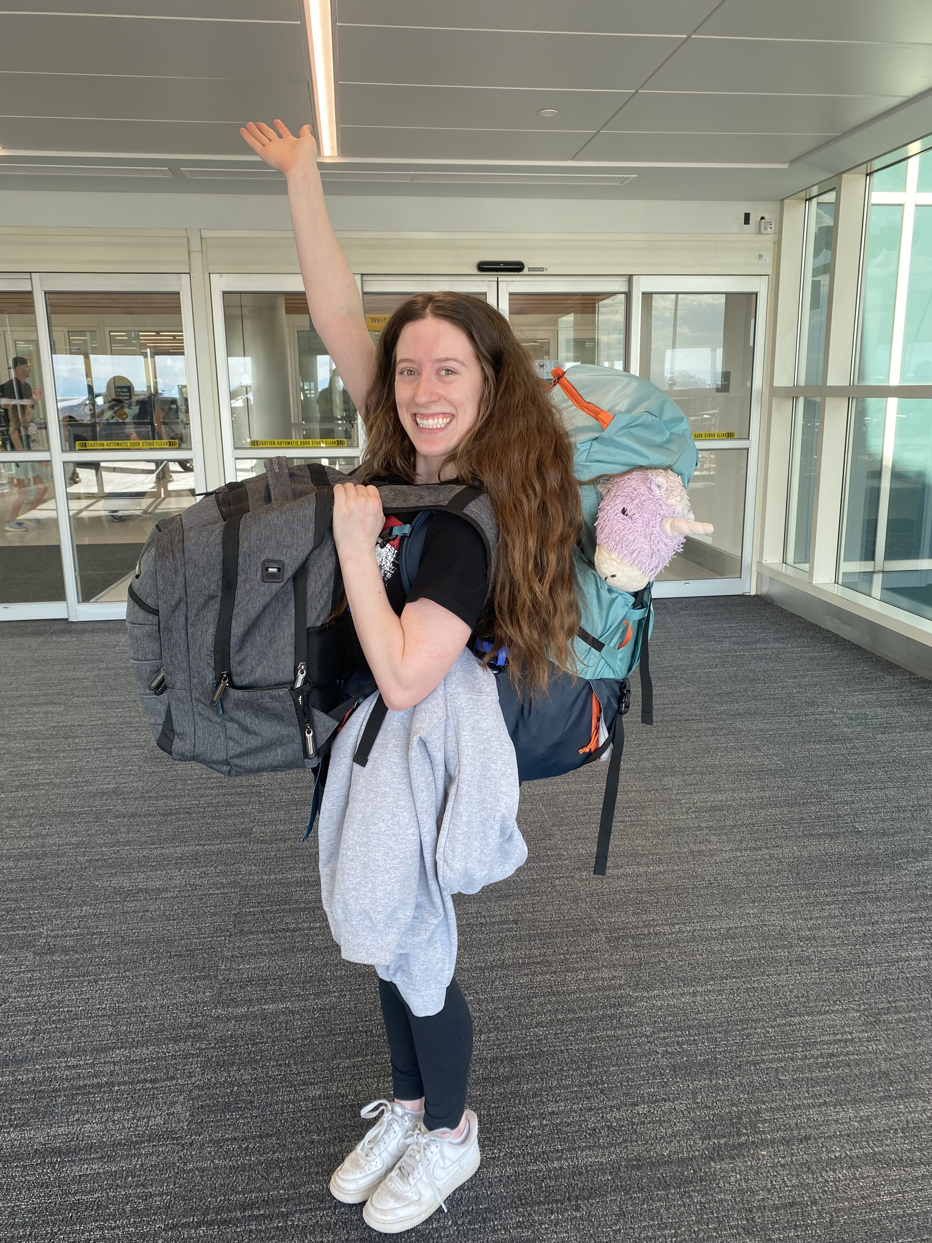 Bella standing at the entrance to the airport with a large 60L backpack on her back and a small grey backpack slung over the front of her body. Her unicorn pillow pet peeks out from the backpack.