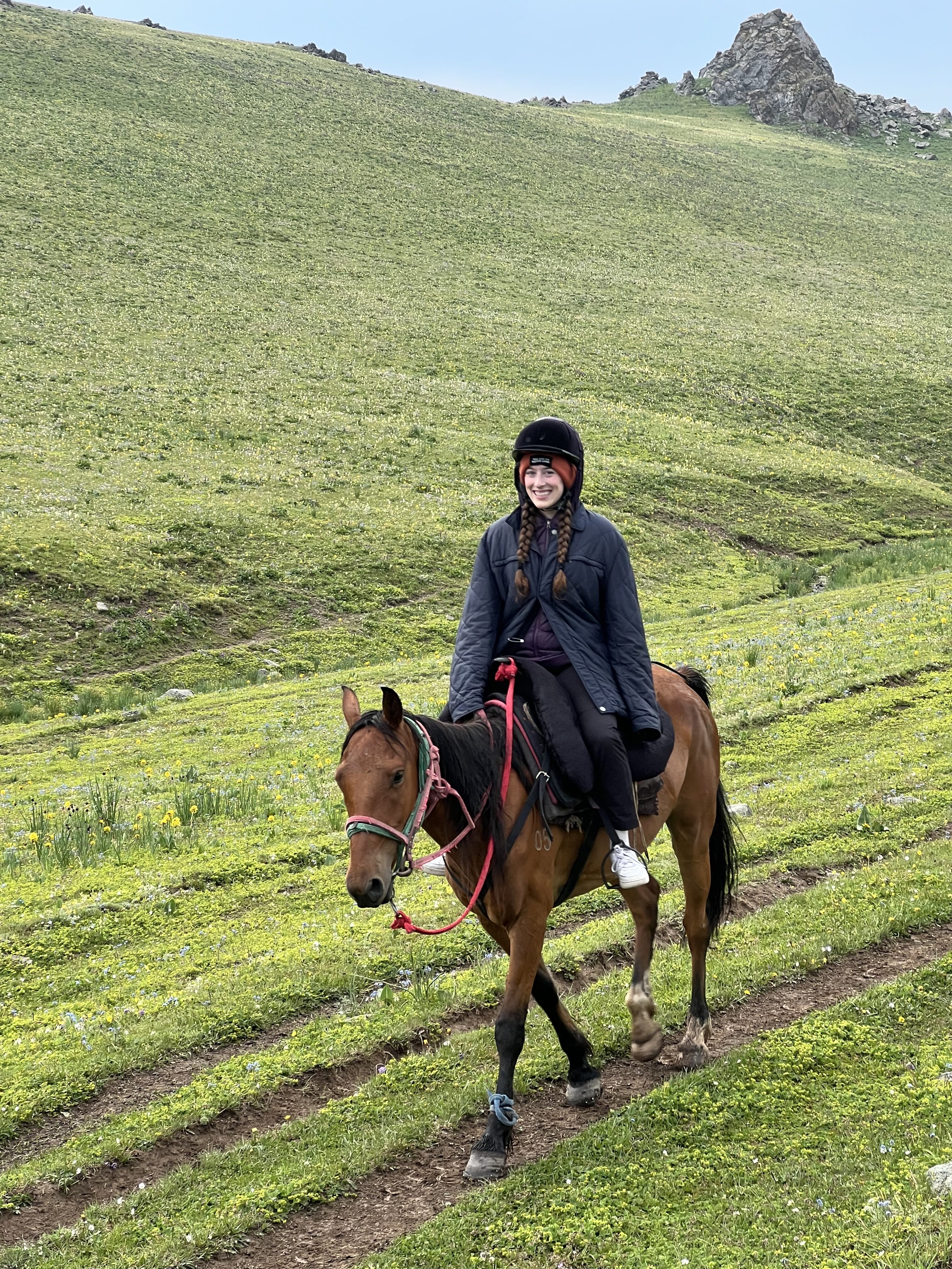 A young woman wearing an oversized navy blue coat and black pants sits on top of a chestnut brown horse with a black mane. The rider is walking on horseback along a vibrant grassy field.