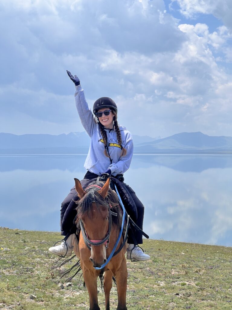A young woman wearing a grey hoodie and blank pants sits on top of a relaxed chestnut brown horse in front of the mirror-like Song Kul lake.