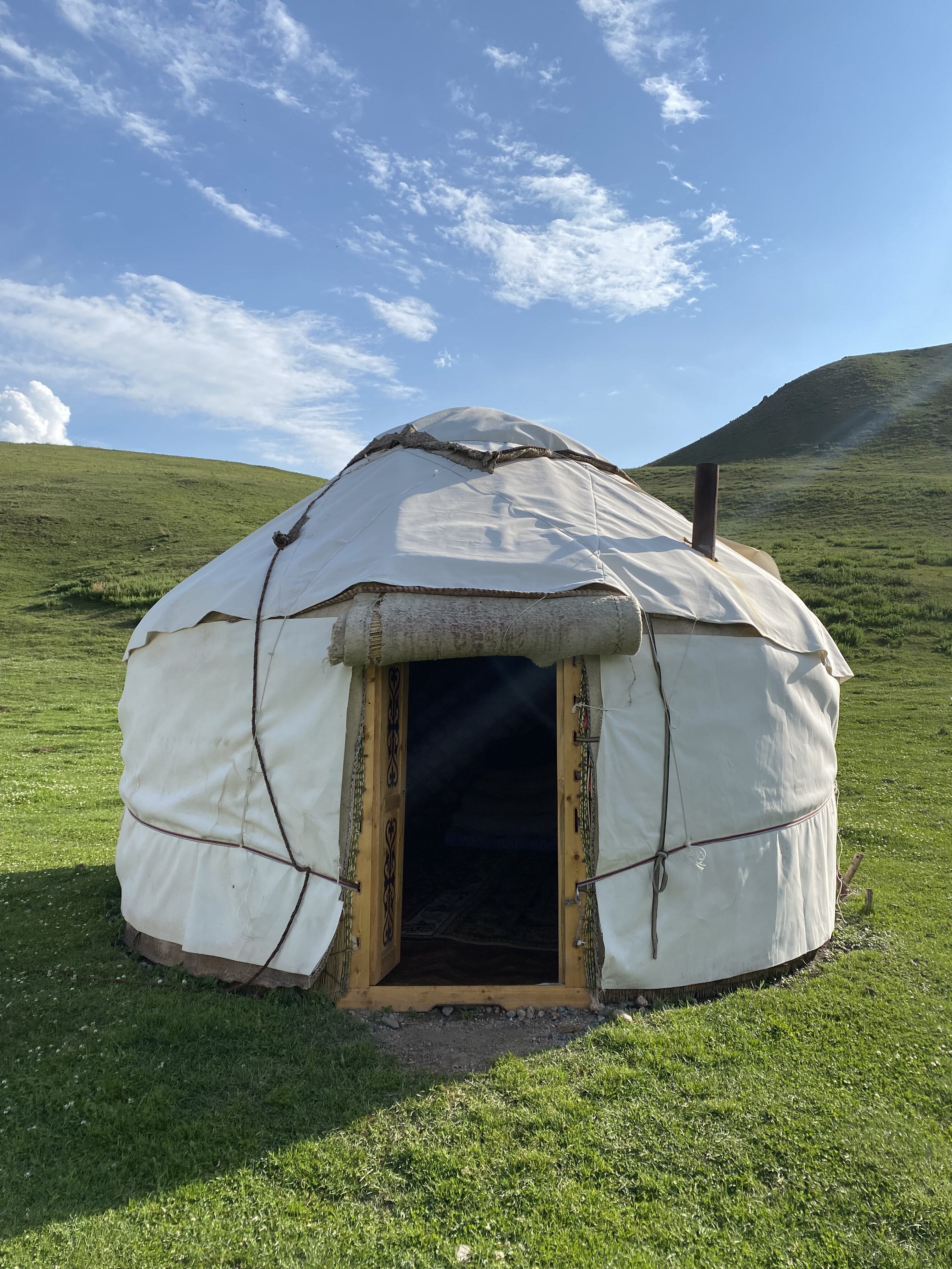 A white yurt with a wooden door frame sitting on the vibrant green grass with a near-cloudless blue sky framing the scene.
