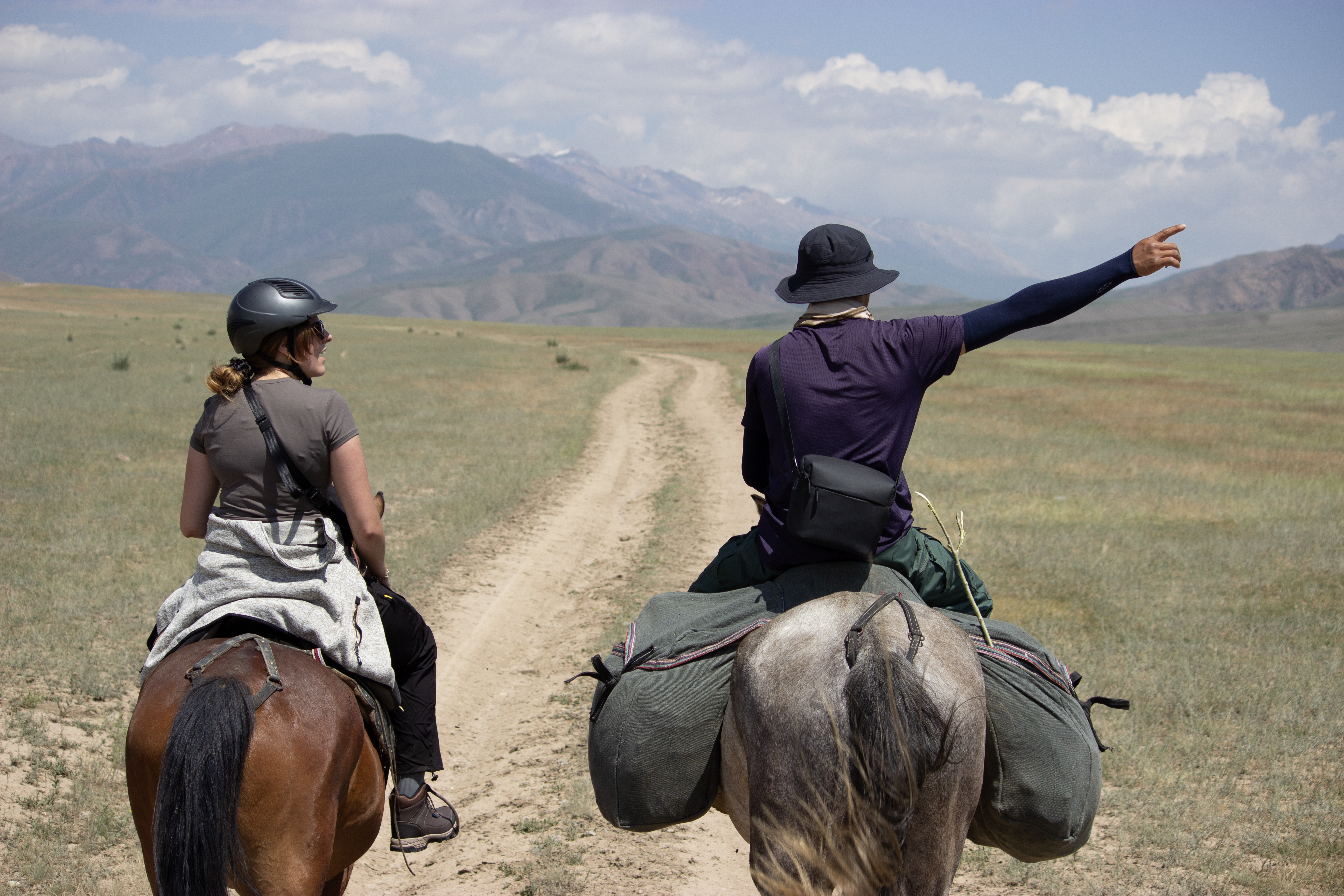 A pair of horseback riders. A young girl rides on a chestnut horse, while the local guide rides a grey horses and points out to the horizon.