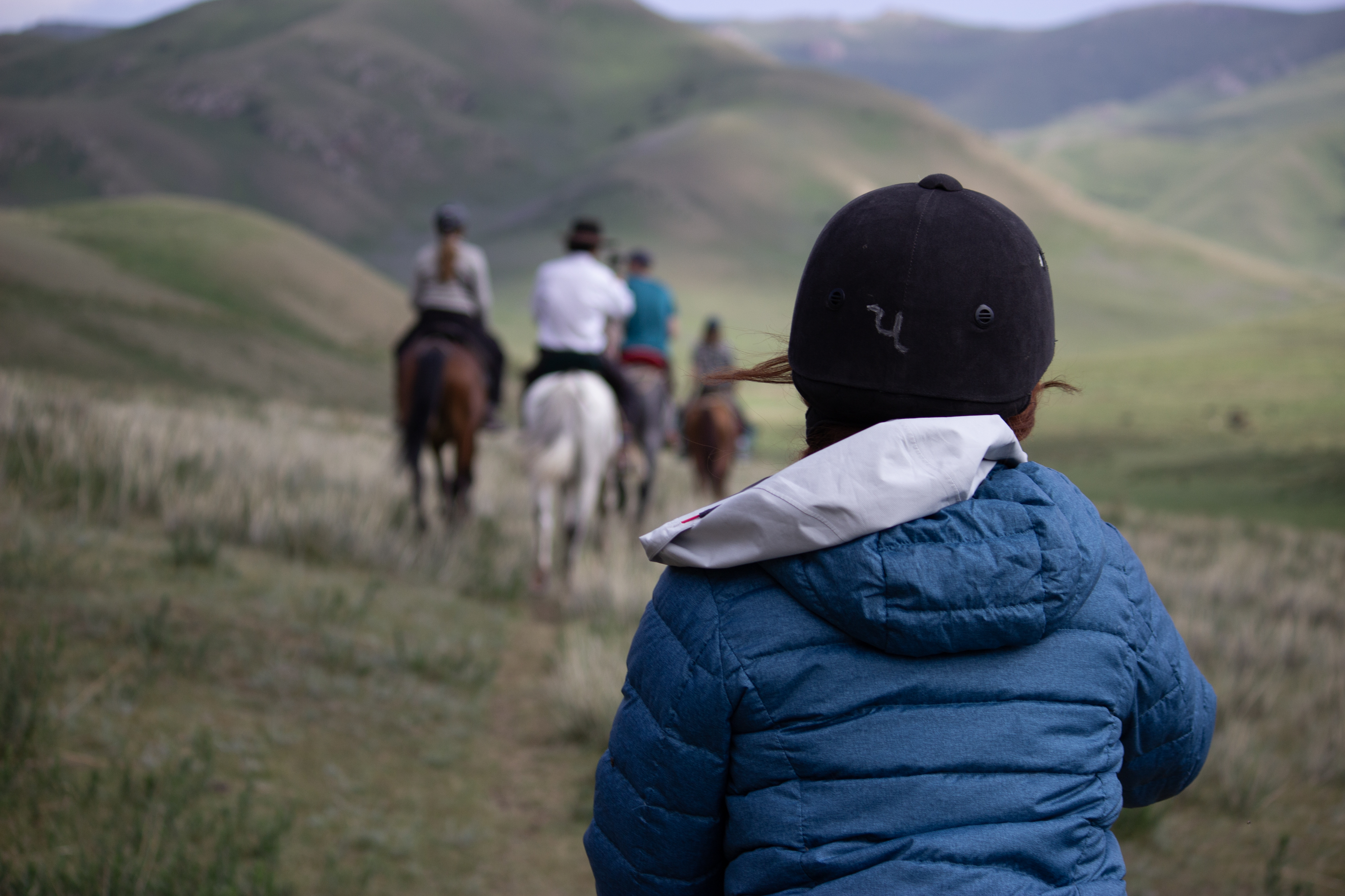 A close-up shot of a young horseback rider wearing a blue coat, with a blurred group of horseback riders in the distance.