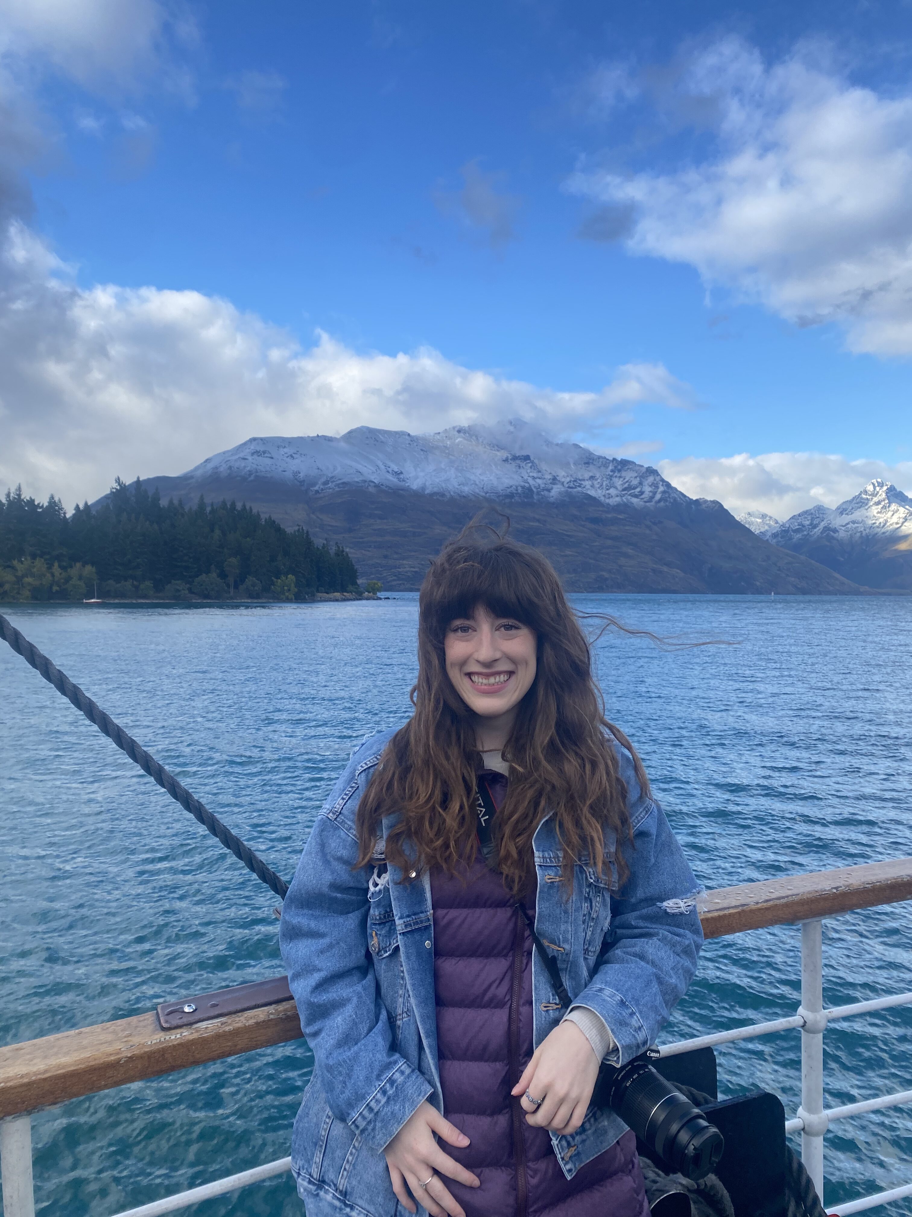 A young woman with brunette bangs wearing a jean jacket over a purple coat smiles on board the TSS Earnslaw in front of Lake Wakatipu and the snowcapped mountains behind her.