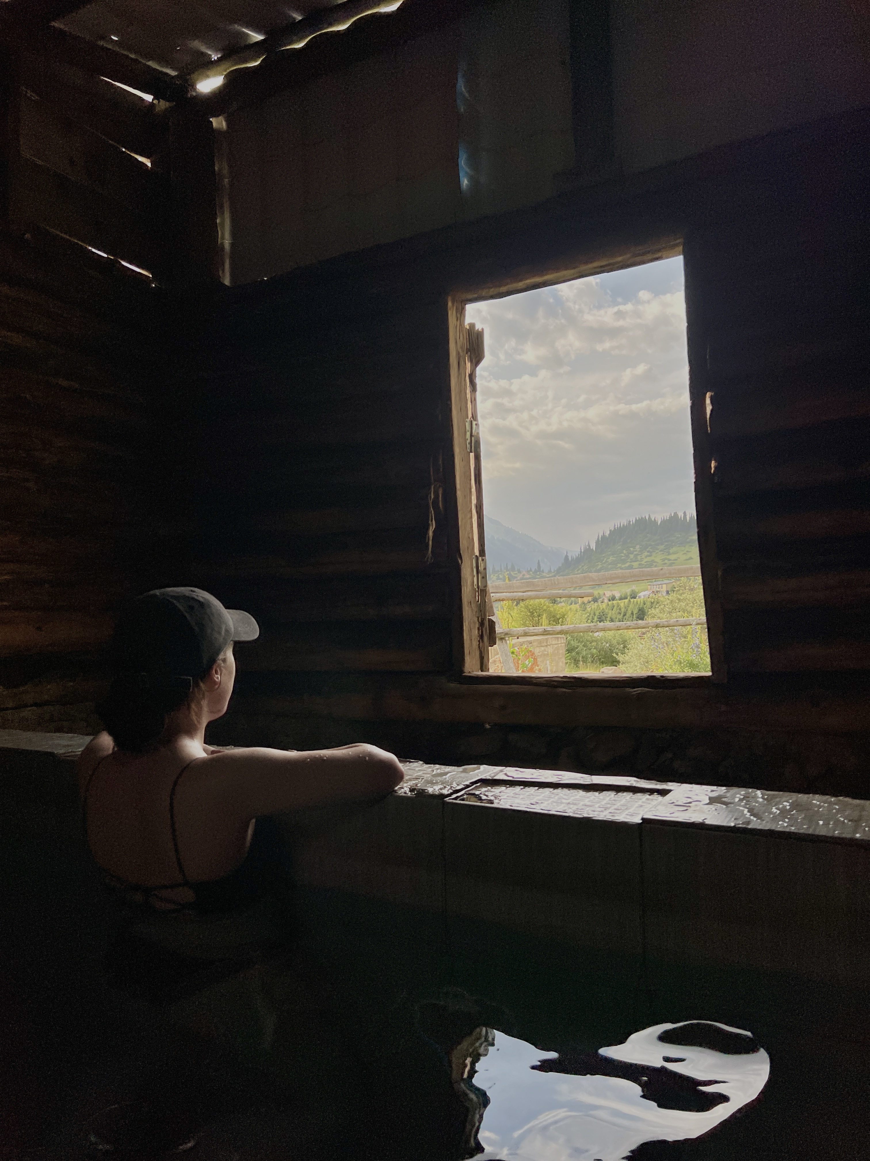 A mostly shadowed young women in an enclosed hot spring wearing a ball cap, perching her arms along a stone railing, gazing out to the light of Altyn Arashan valley.