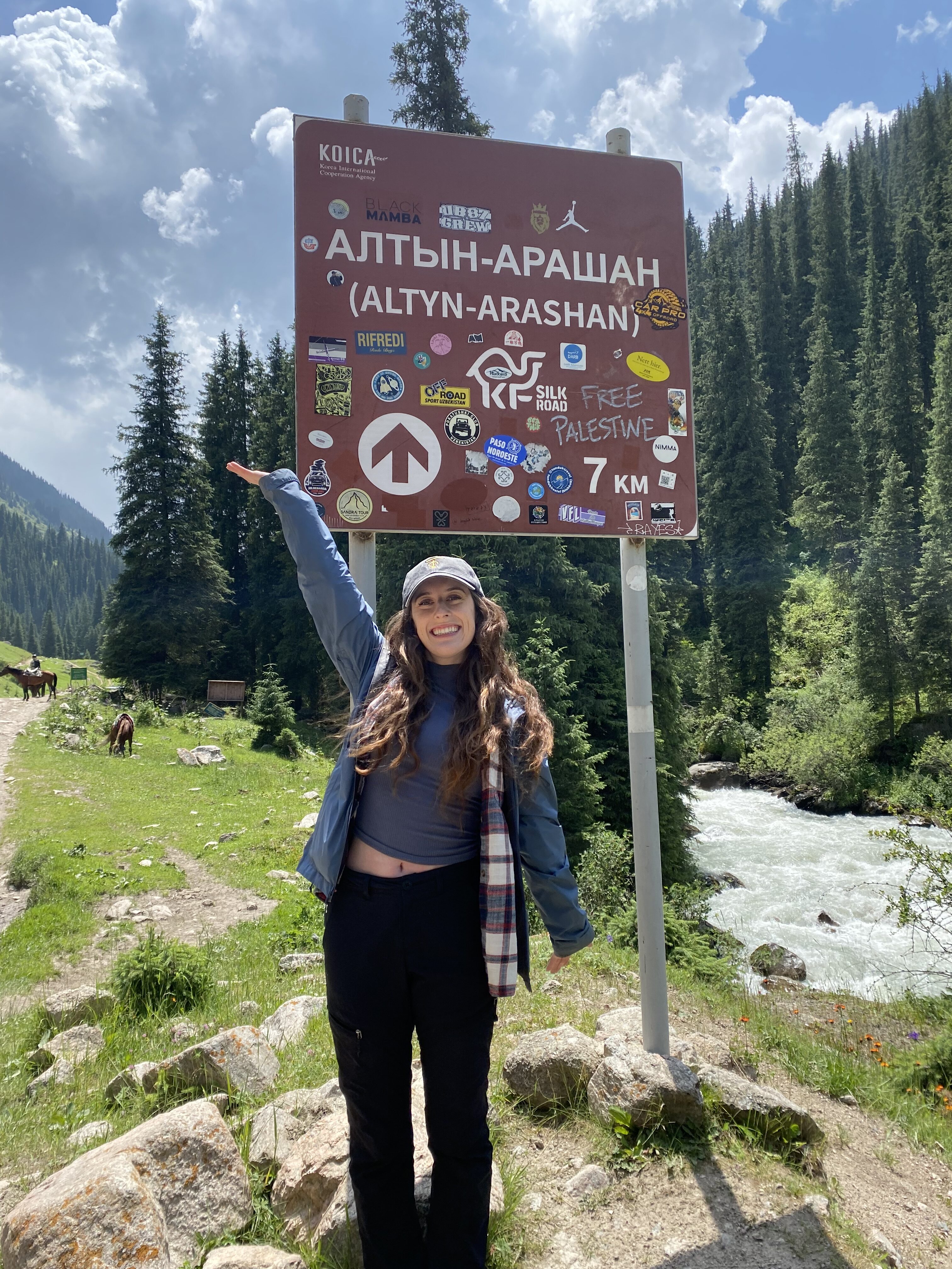 A young women wearing a blue rain coat with a grey cap and blank pants posing under a brown sign decorated with lots of stickers that says "Altyn Arashan" in Kyrgyz and English.