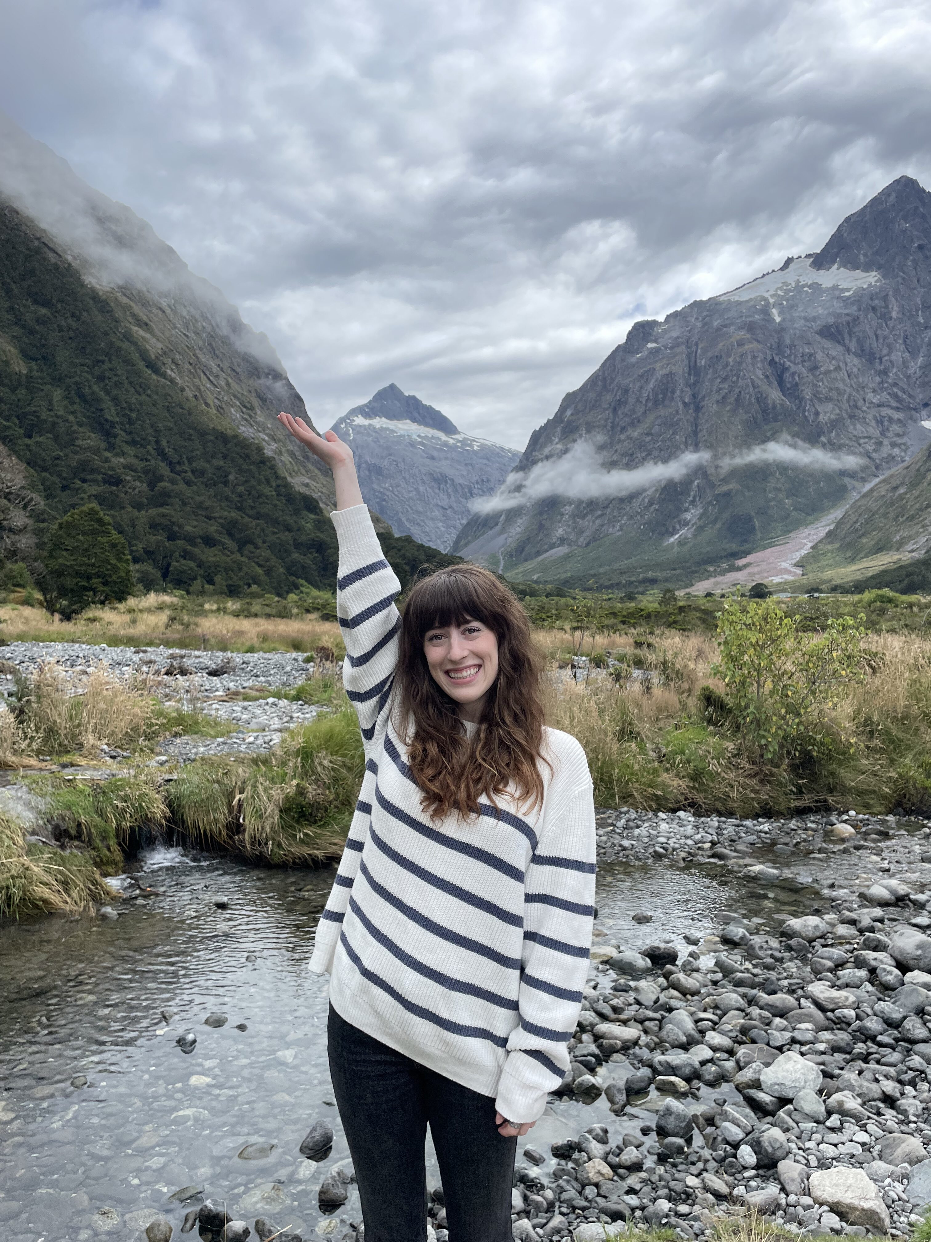 A young woman with brunette bangs wearing a striped white sweater poses with her arm up in front of a flowing stream and the mountains outside Fiordland National Park.