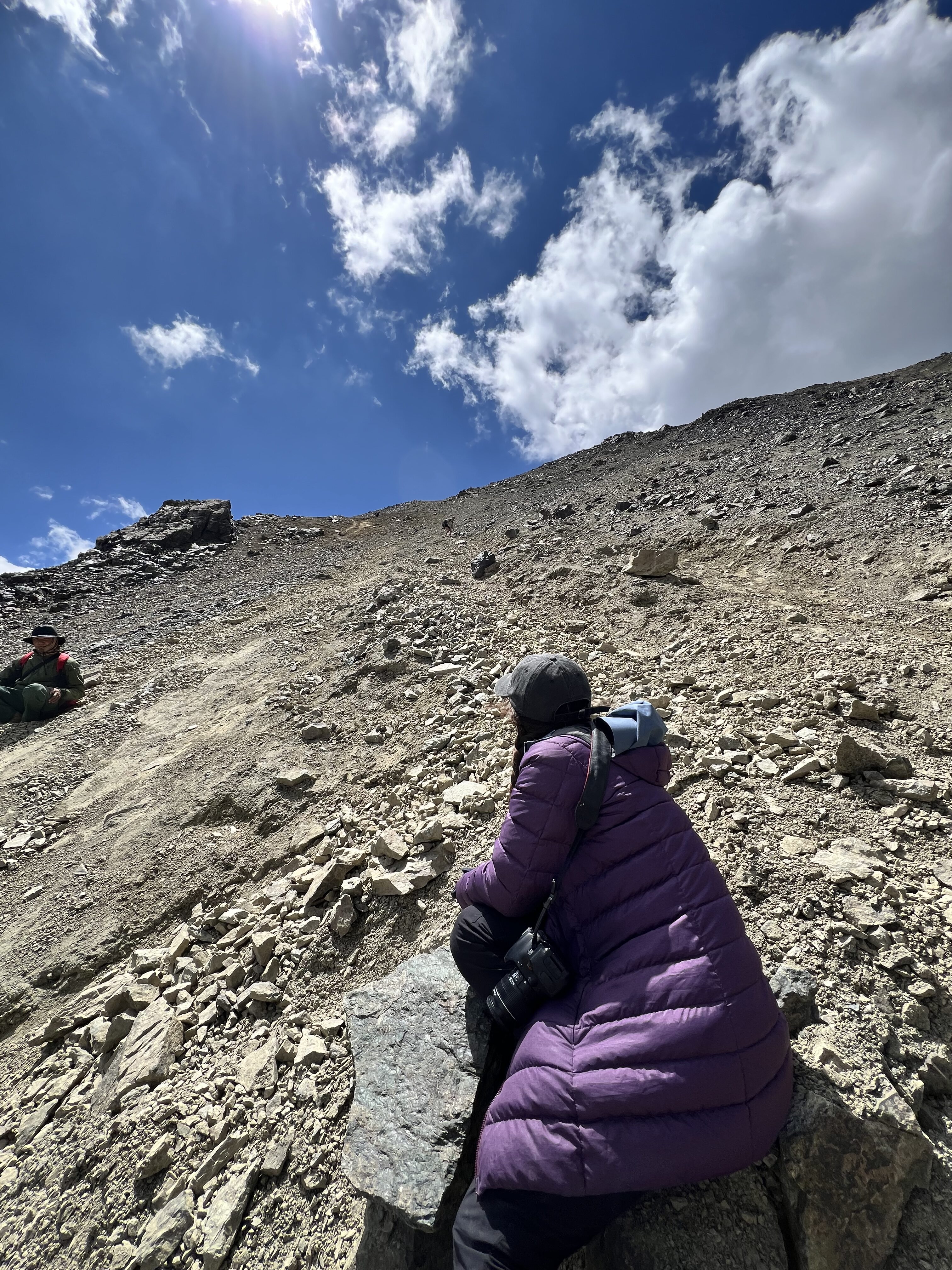 A young woman wearing a purple coat sits along the steep gravel trail of Ala Kul pass, gazing at the rest of the trek above her.
