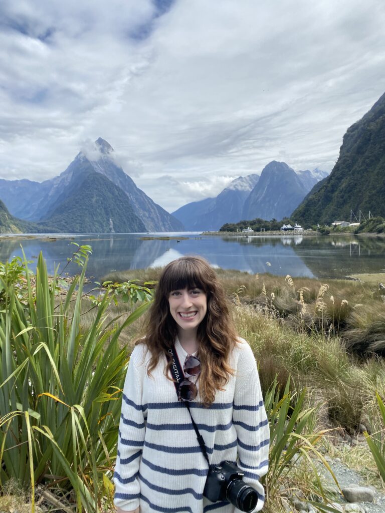 A brunette woman with bangs and a white striped sweater with a camera slung around her body smiles in front of Milford Sound.