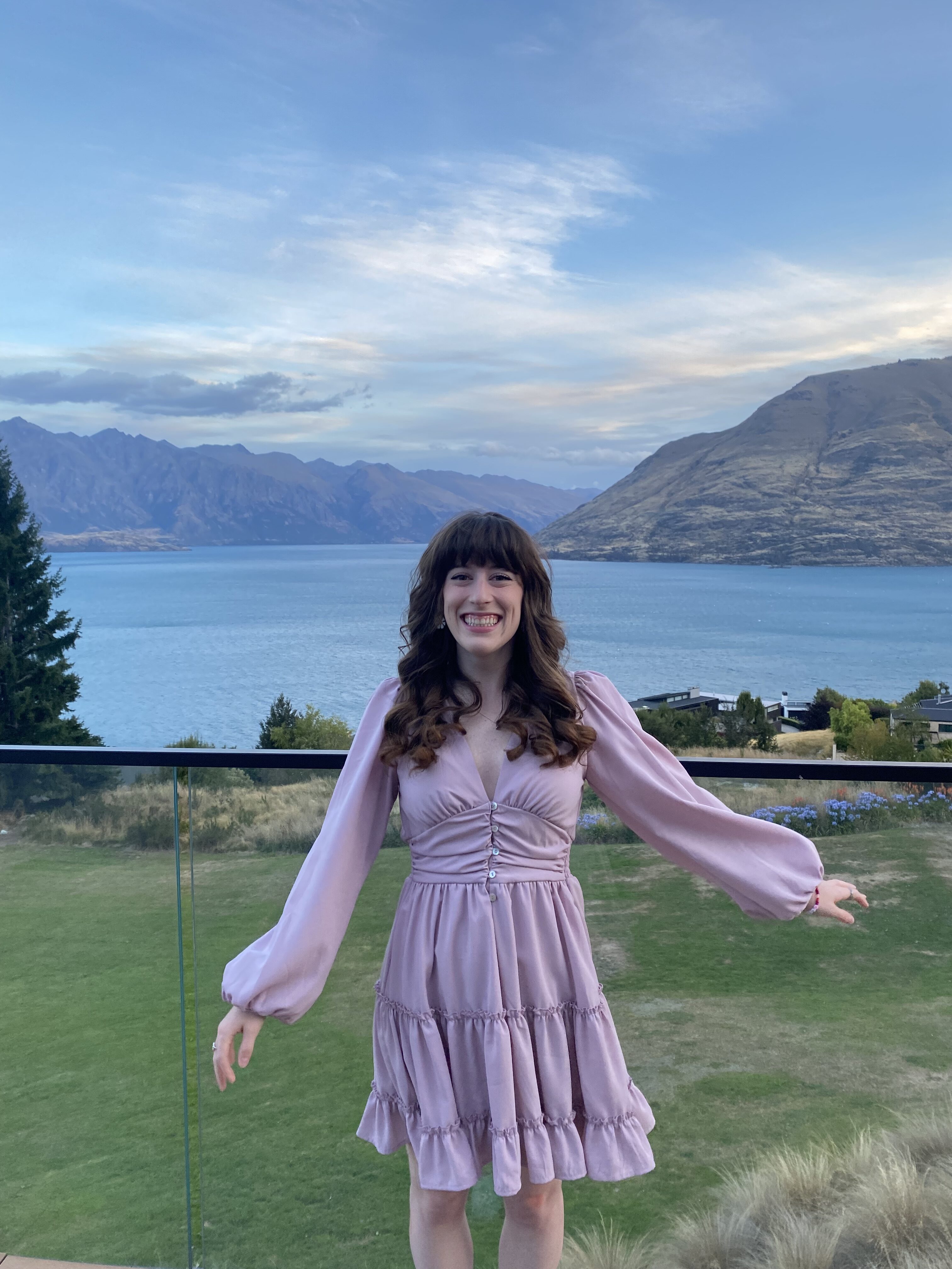 A young woman with curled hair and bangs wearing a pink dress, posing in front of Lake Wakatipu and the mountains in the background.