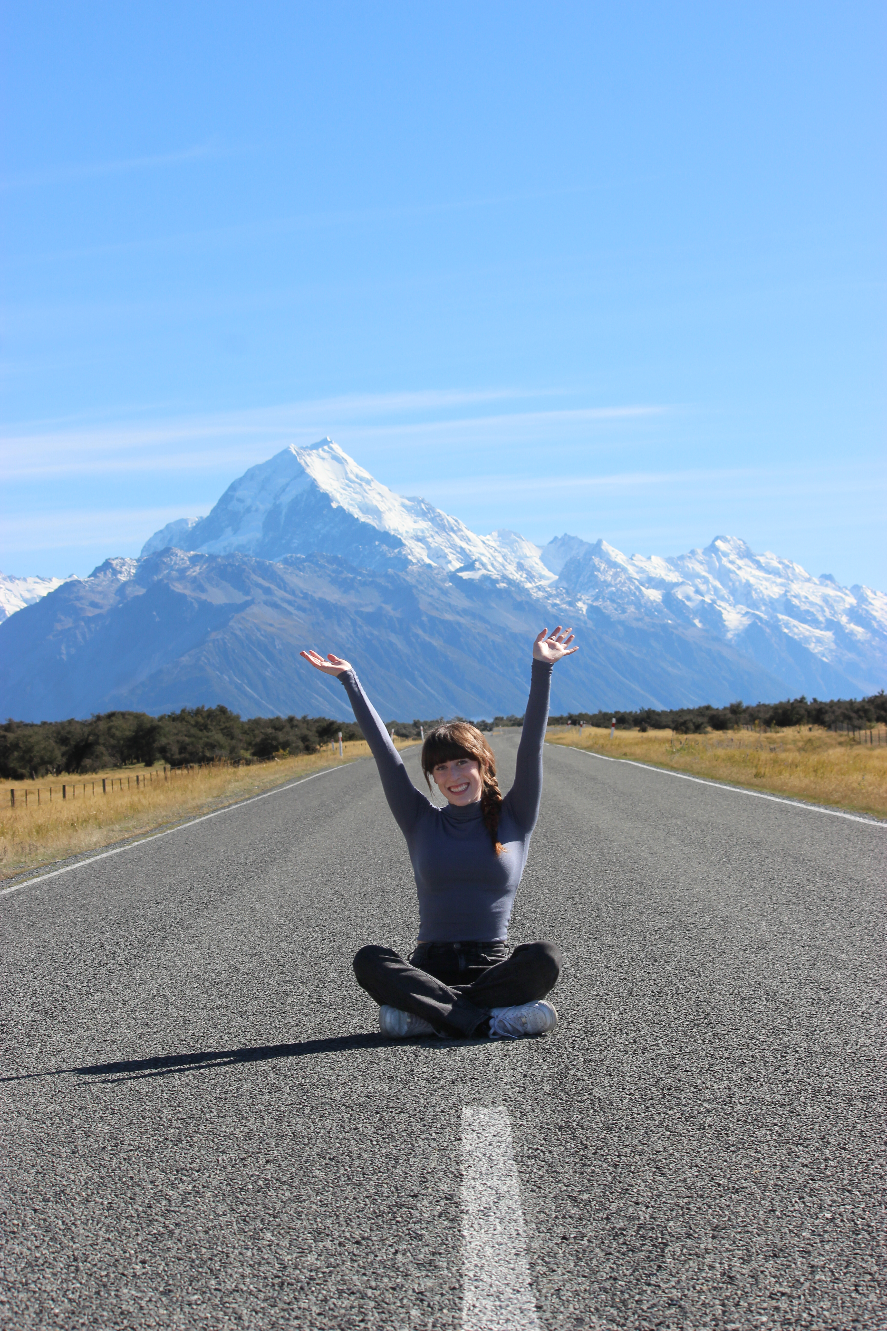 A young woman sitting criss-cross with her arms in the air, in the middle of the road in Aoraki/Mt. Cook National Park. The mountain can be seen in the background.
