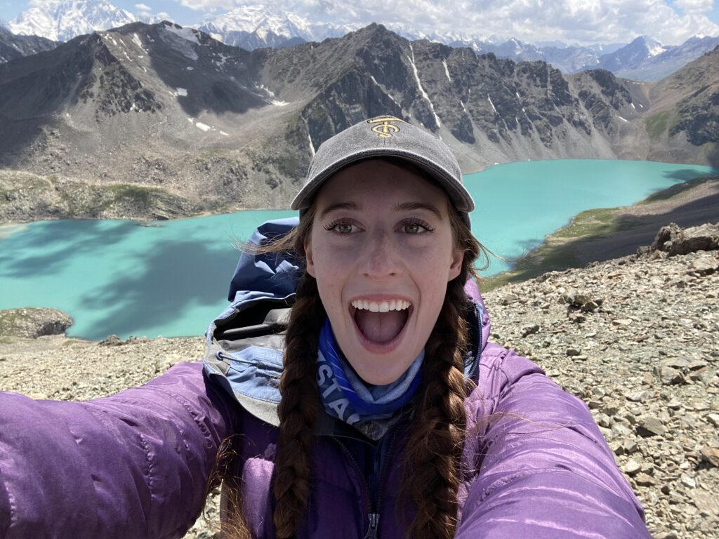 A young women wearing two braids, a grey ball cap, and a purple coat, poses in awe in front of Ala Kul Lake.