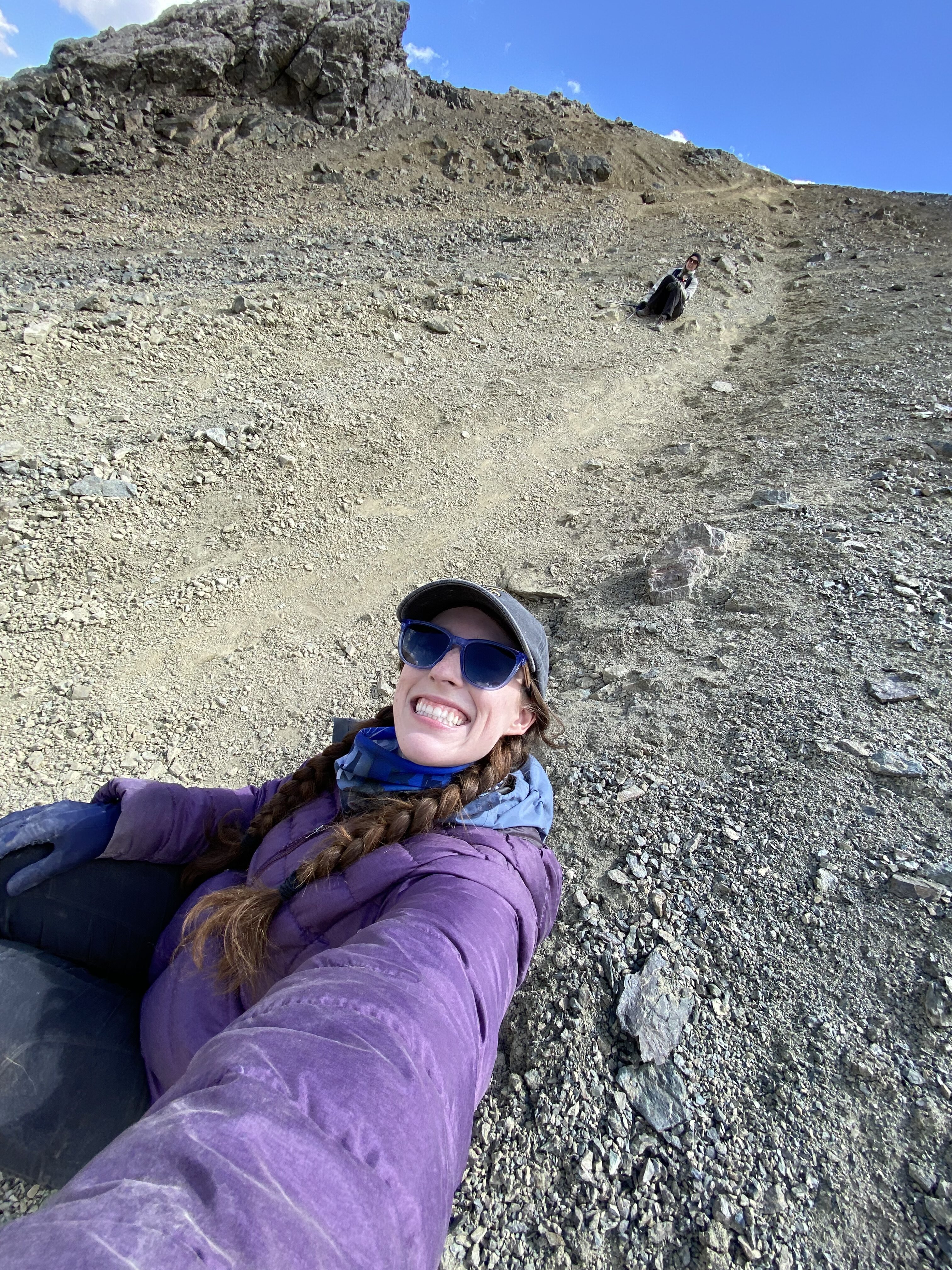 A young women with two braids, a purple coat and a grey baseball cap taking a selfie along the gravel trail of Ala Kul Pass. Behind her is another young hiker.
