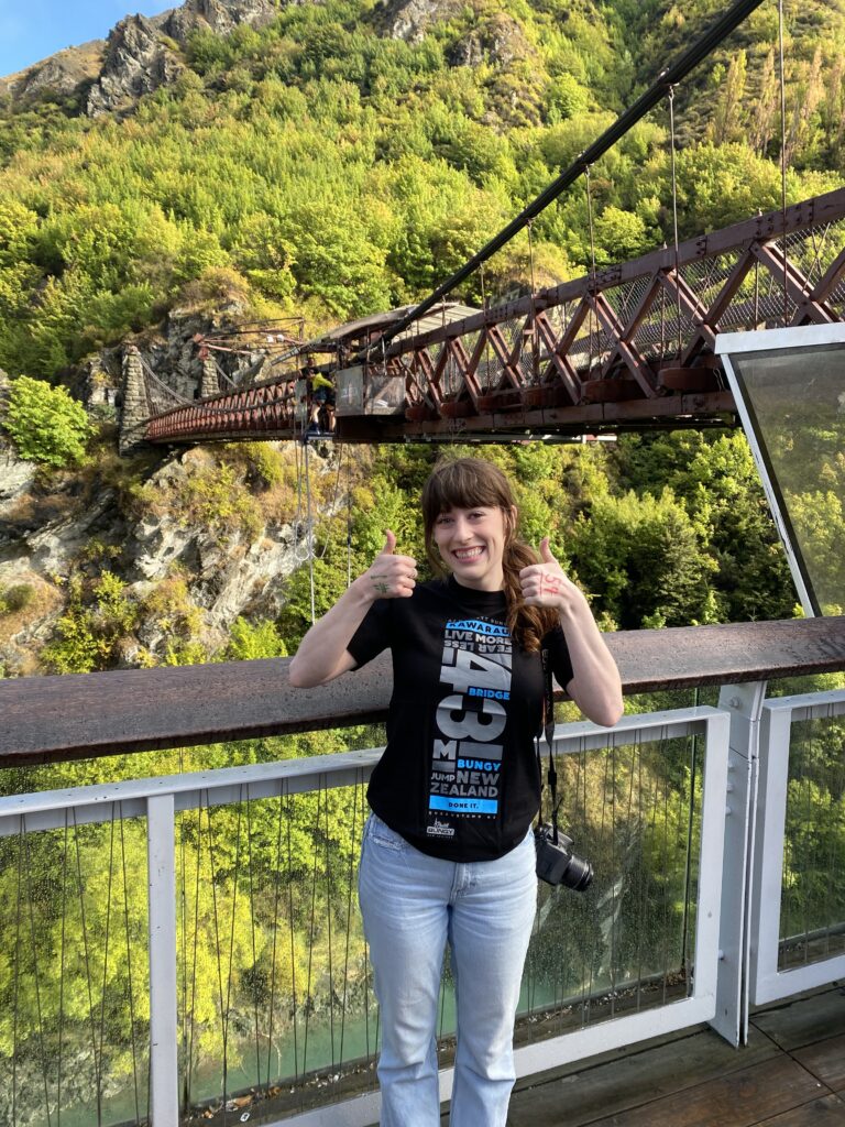 A daring adventuring posing with two thumbs up in front of Kawarau Bridge, the site of the first bungee jump in New Zealand.