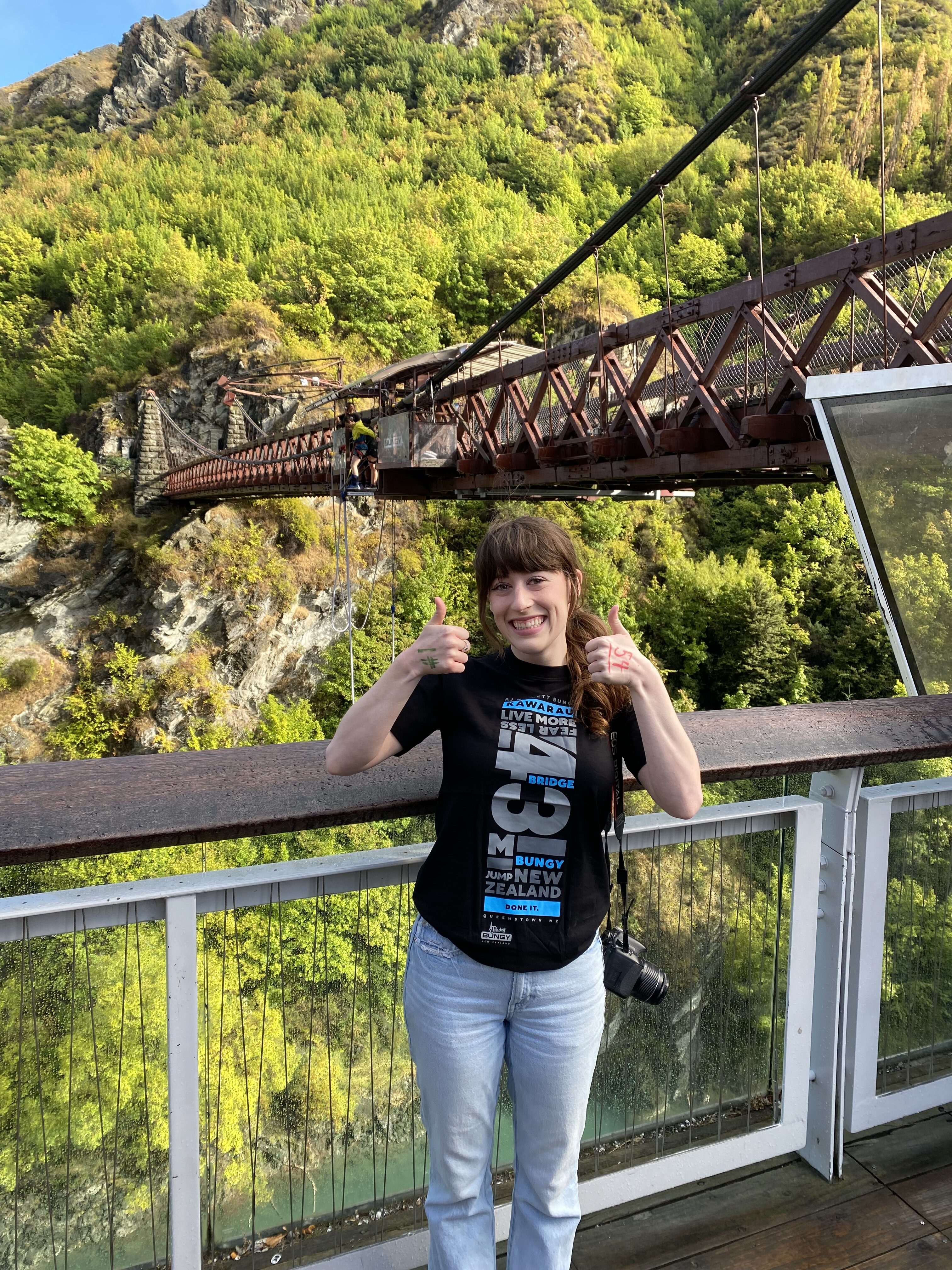 A daring adventuring posing with two thumbs up in front of Kawarau Bridge, the site of the first bungee jump in New Zealand.