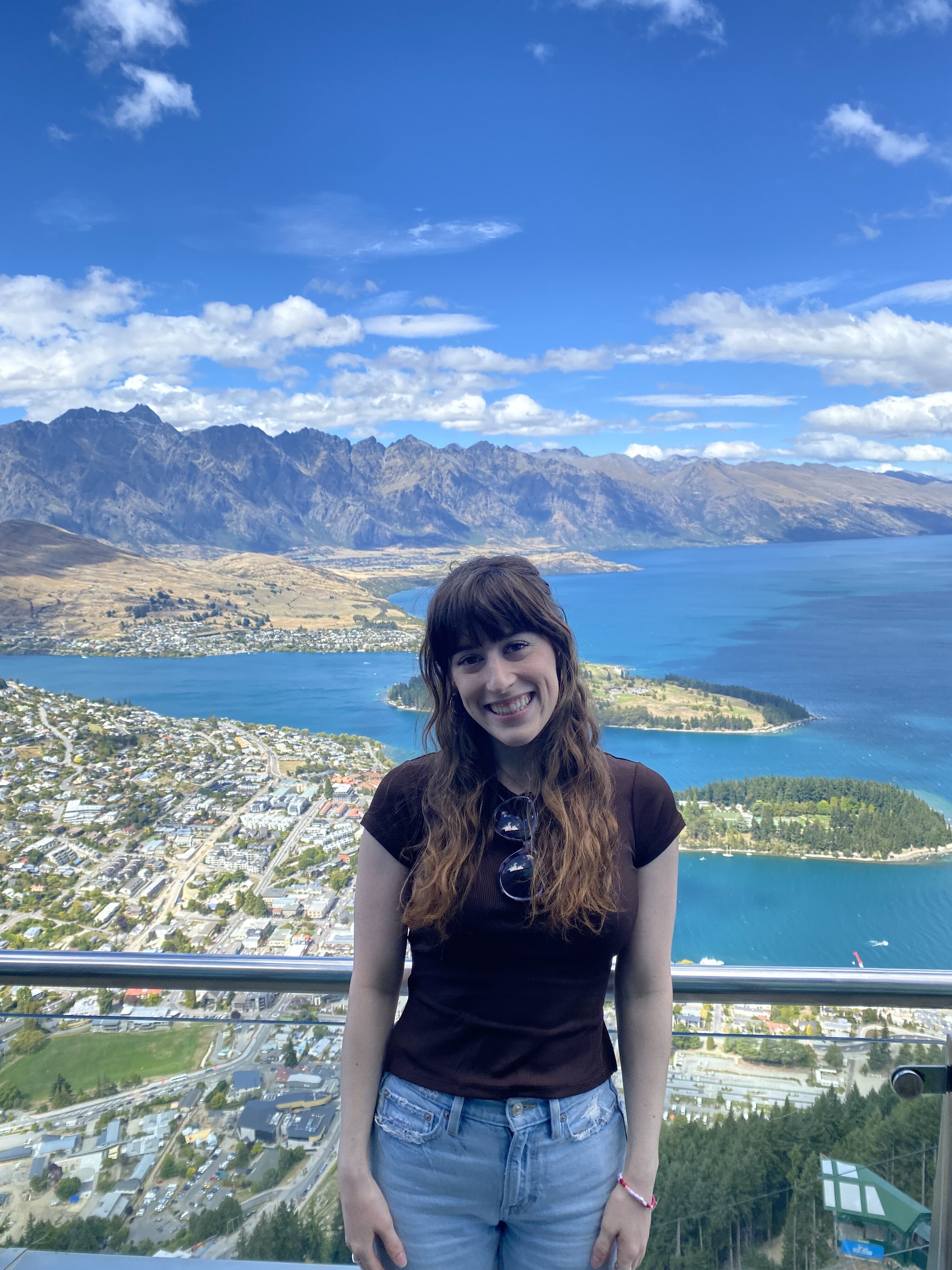 A young woman with bangs wearing a brown shirt and light denim jeans smiles in front of the city of Queenstown and The Remarkables at the top of the Queenstown Skyline Gondola.