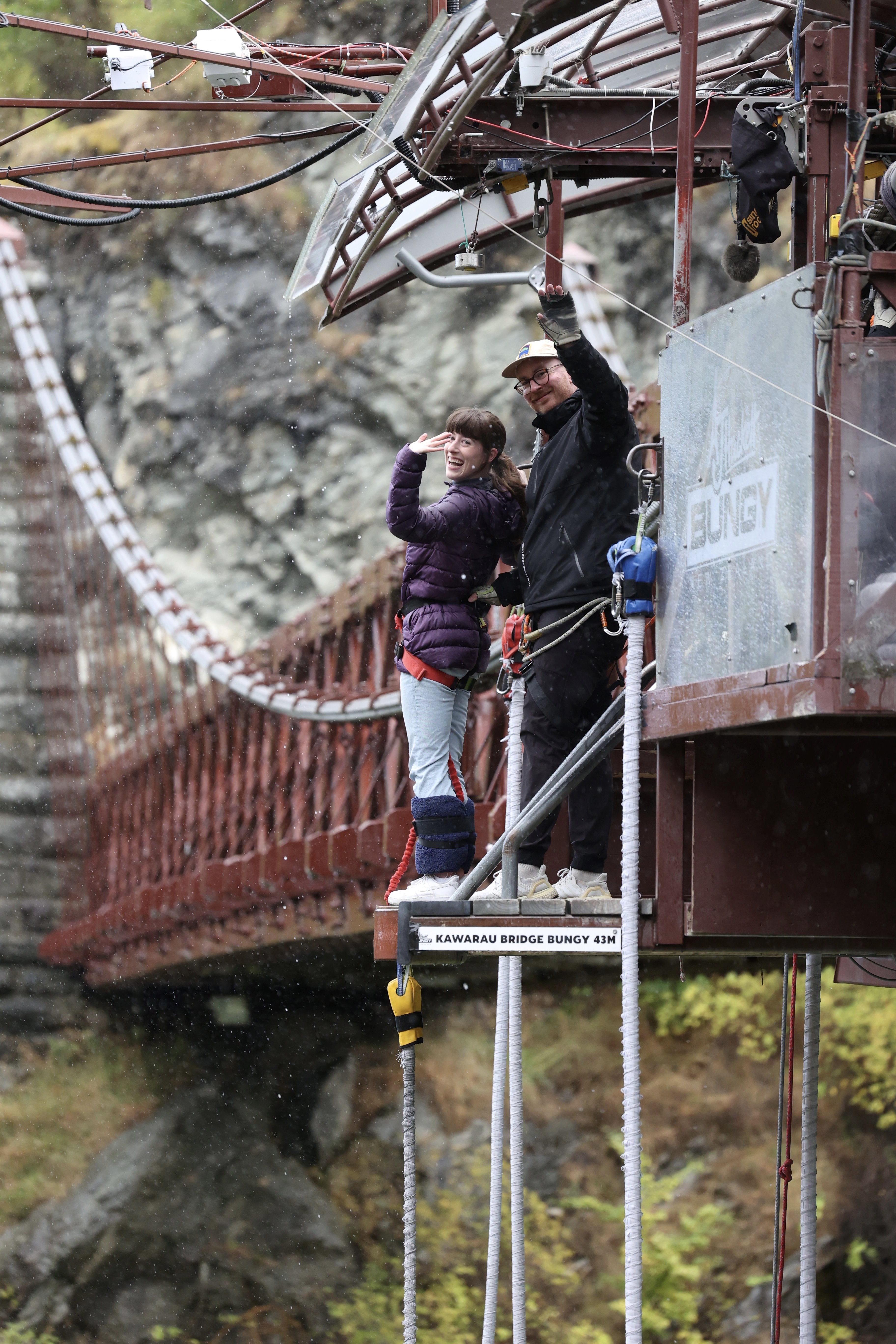 A young woman wearing a purple coat and light blue denim waving from the top of the bungee platform with her bungee guide at Kawarau bridge – a famous bungee jumping spot in Queenstown.