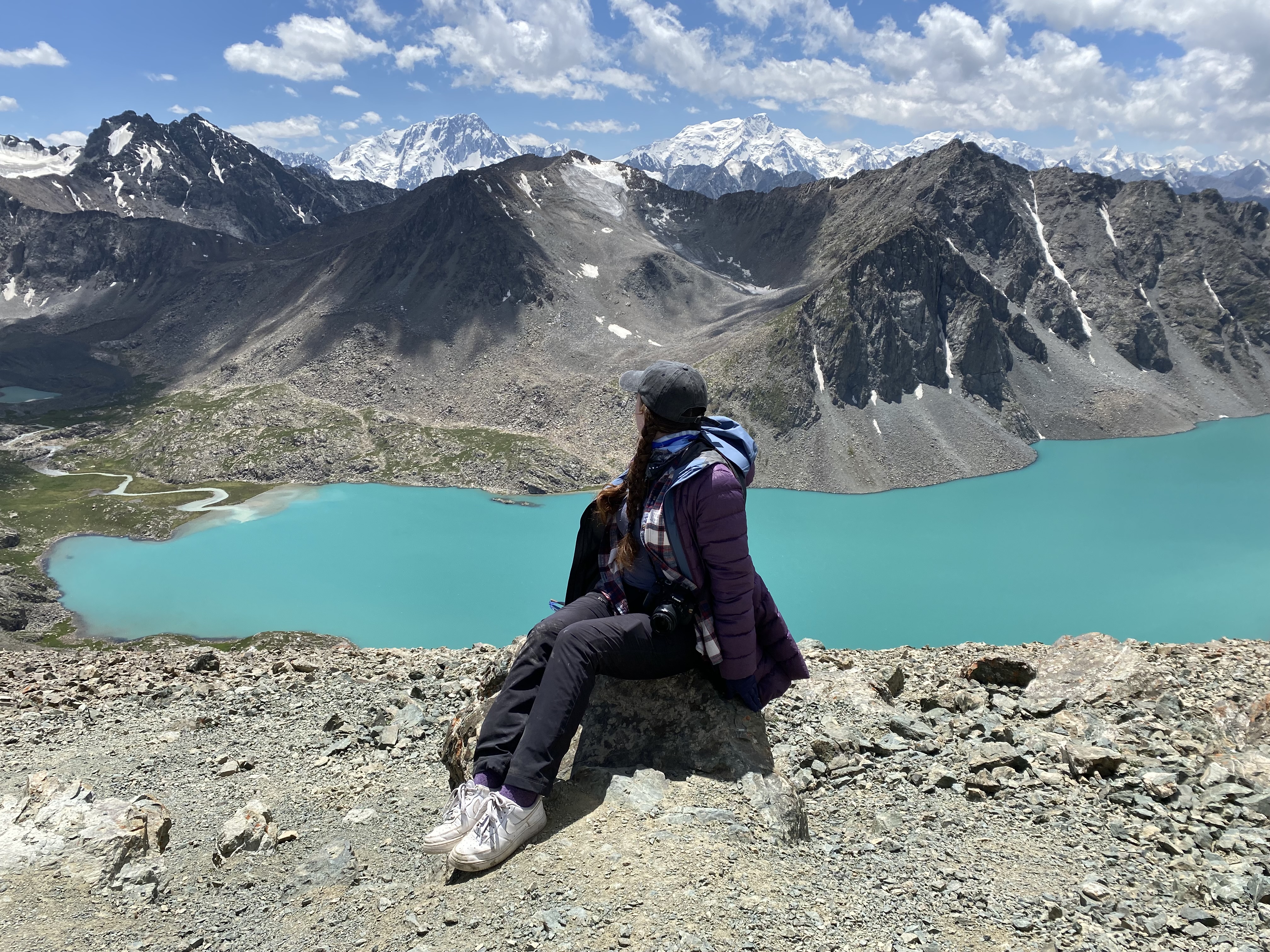 A young women wearing a purple coat, grey ball cap, black pants and worn white tennis shoes, gazes upon a turquoise blue lake – Ala Kul Lake – nestled between rocky, grey mountains in Kyrgyzstan.