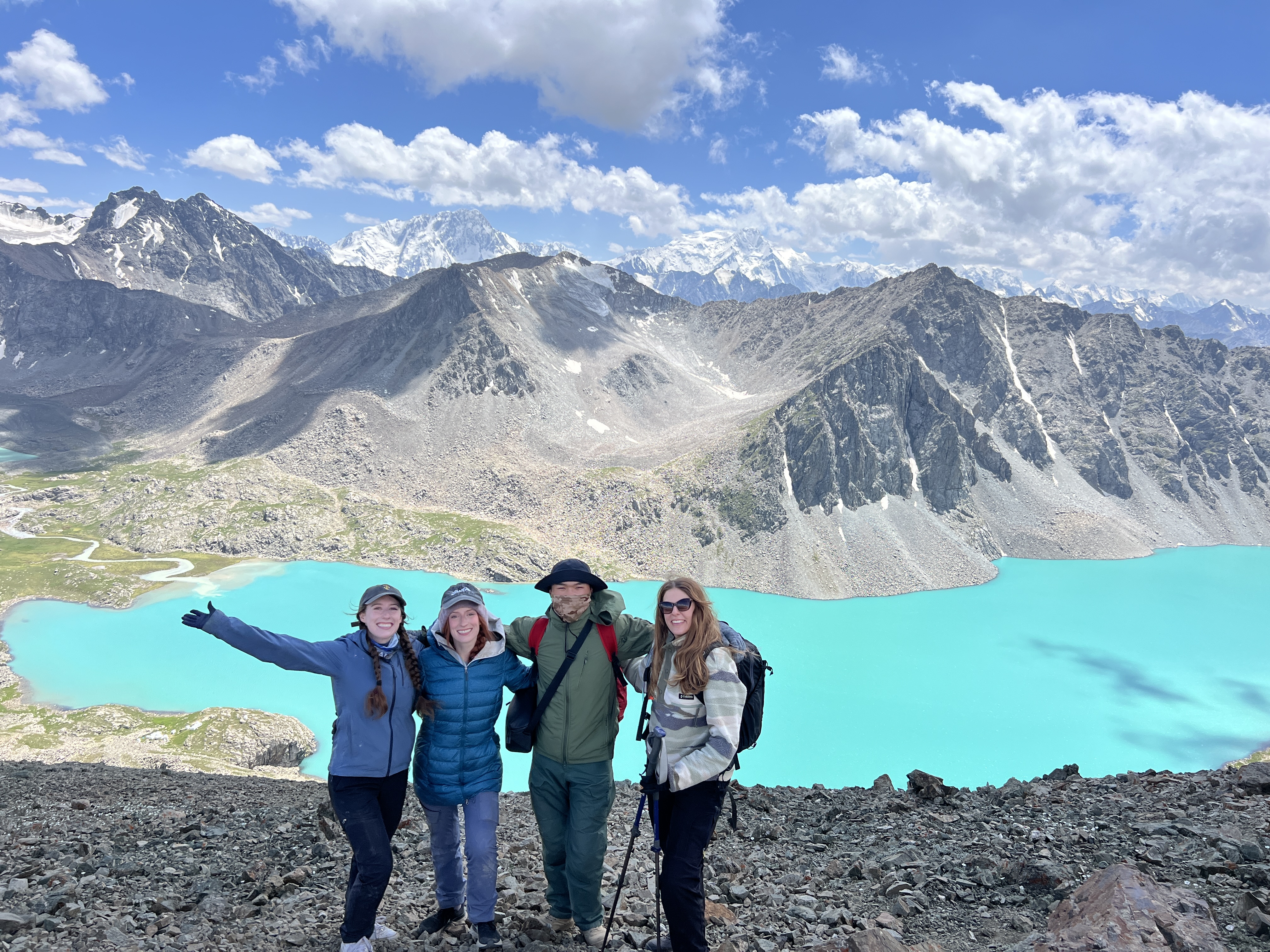 A group of four hikers at the top of Ala Kul pass, smiling with the turquoise lake in the background, surrounded by jagged grey mountains and a cloud-filled blue sky.