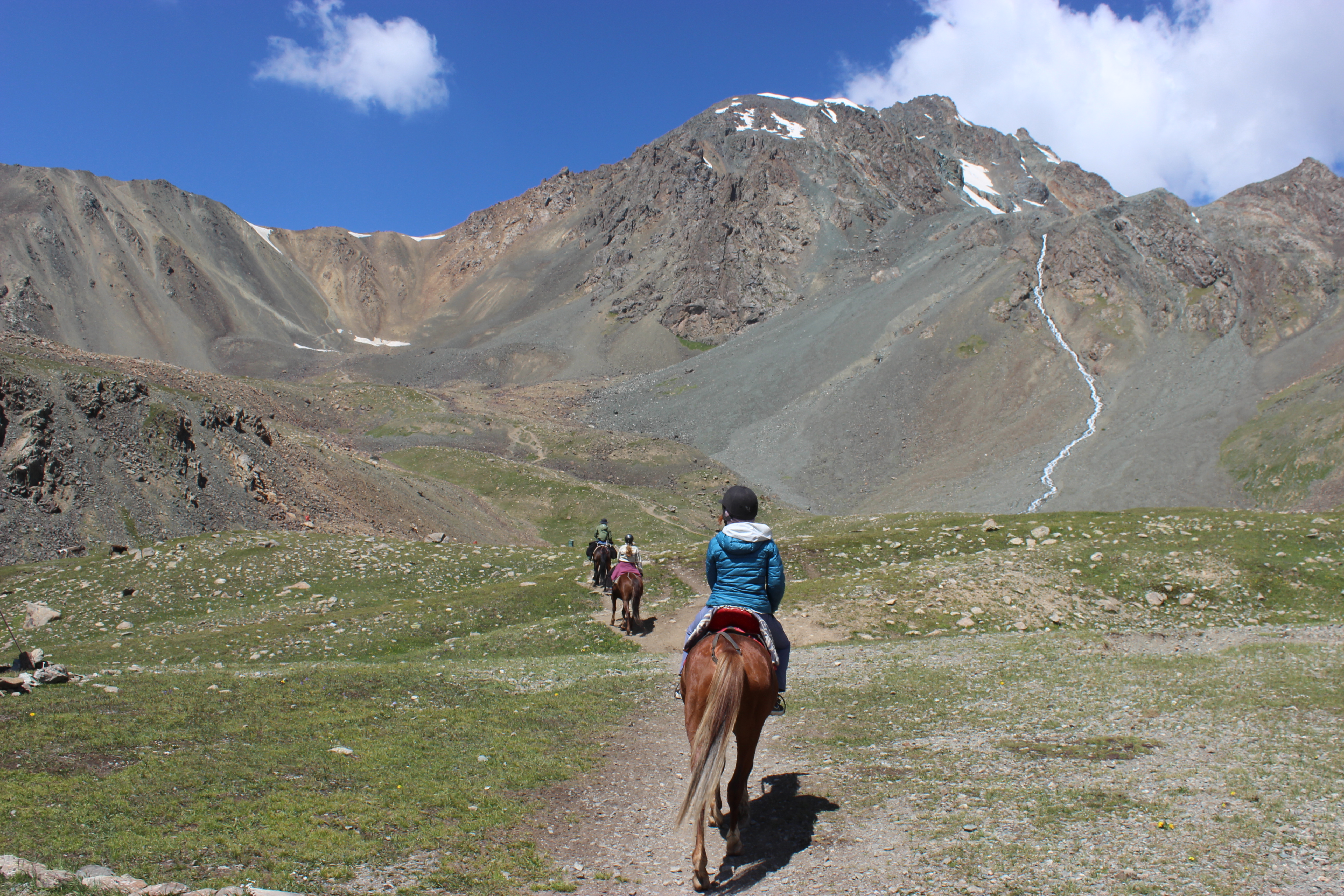 A small group of horseback riders riding towards a bare, rock-covered Mountain in Kyrgyzstan. The base of this mountain is Keldike Gorge.