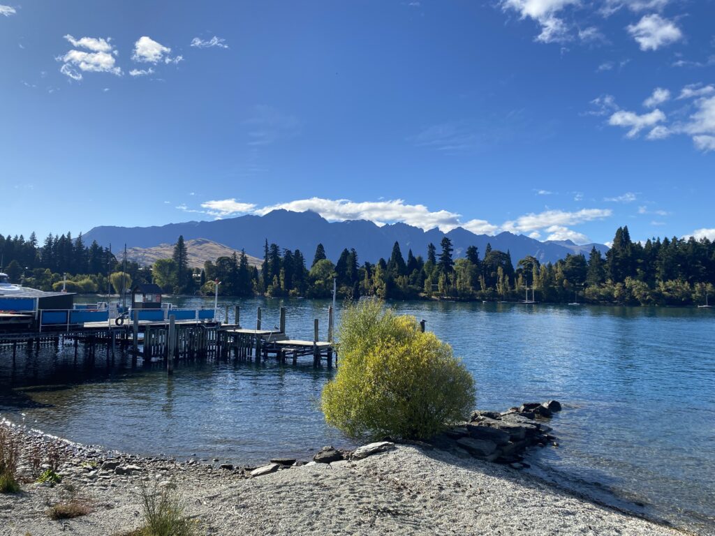 A dock on Lake Wakatipu with The Remarkable peaking through the trees in the background.