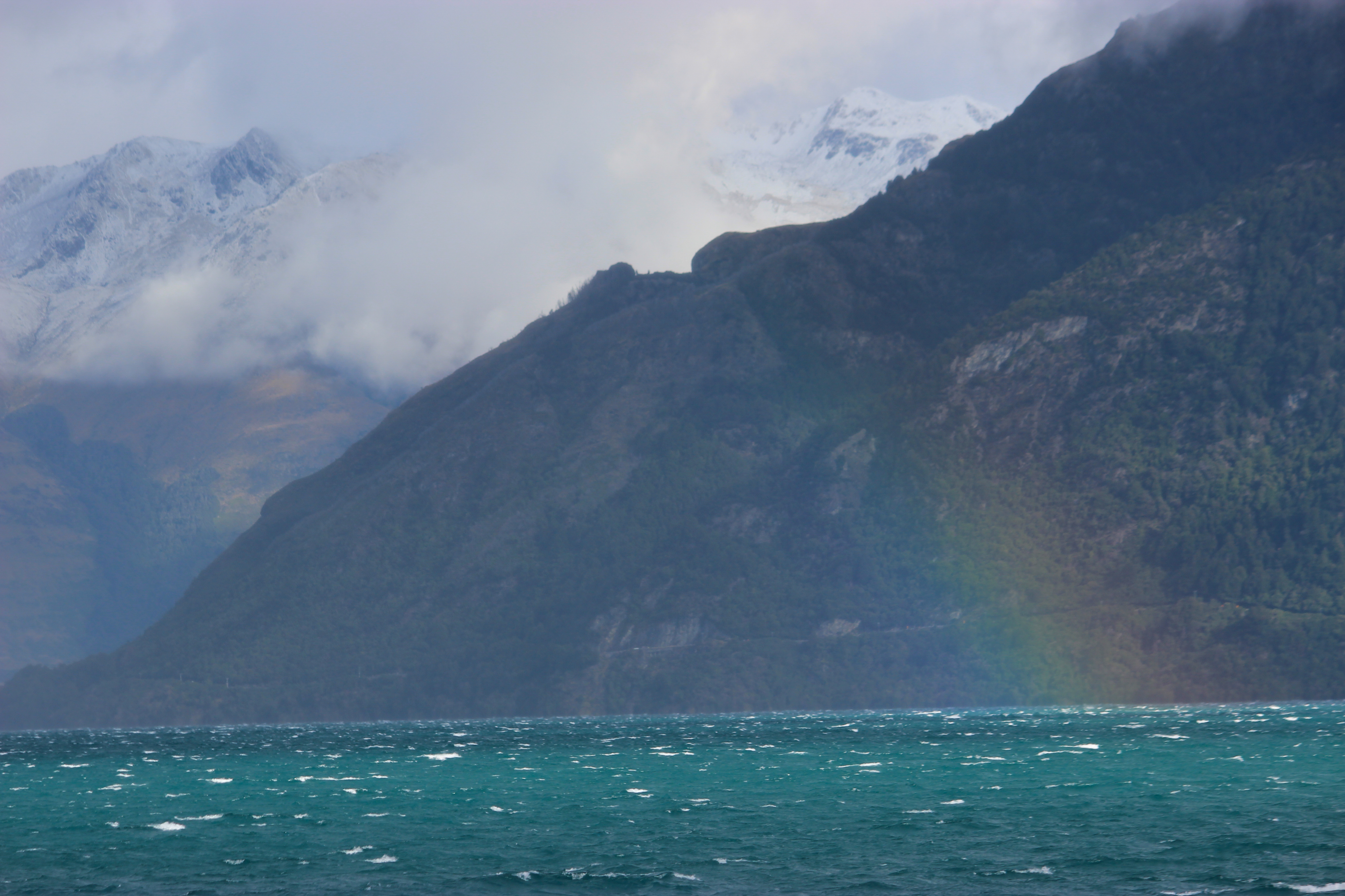 A beautiful rainbow over Lake Wakatipu in Queenstown with a dark blue mountain behind it and the turquoise lake water beneath it.