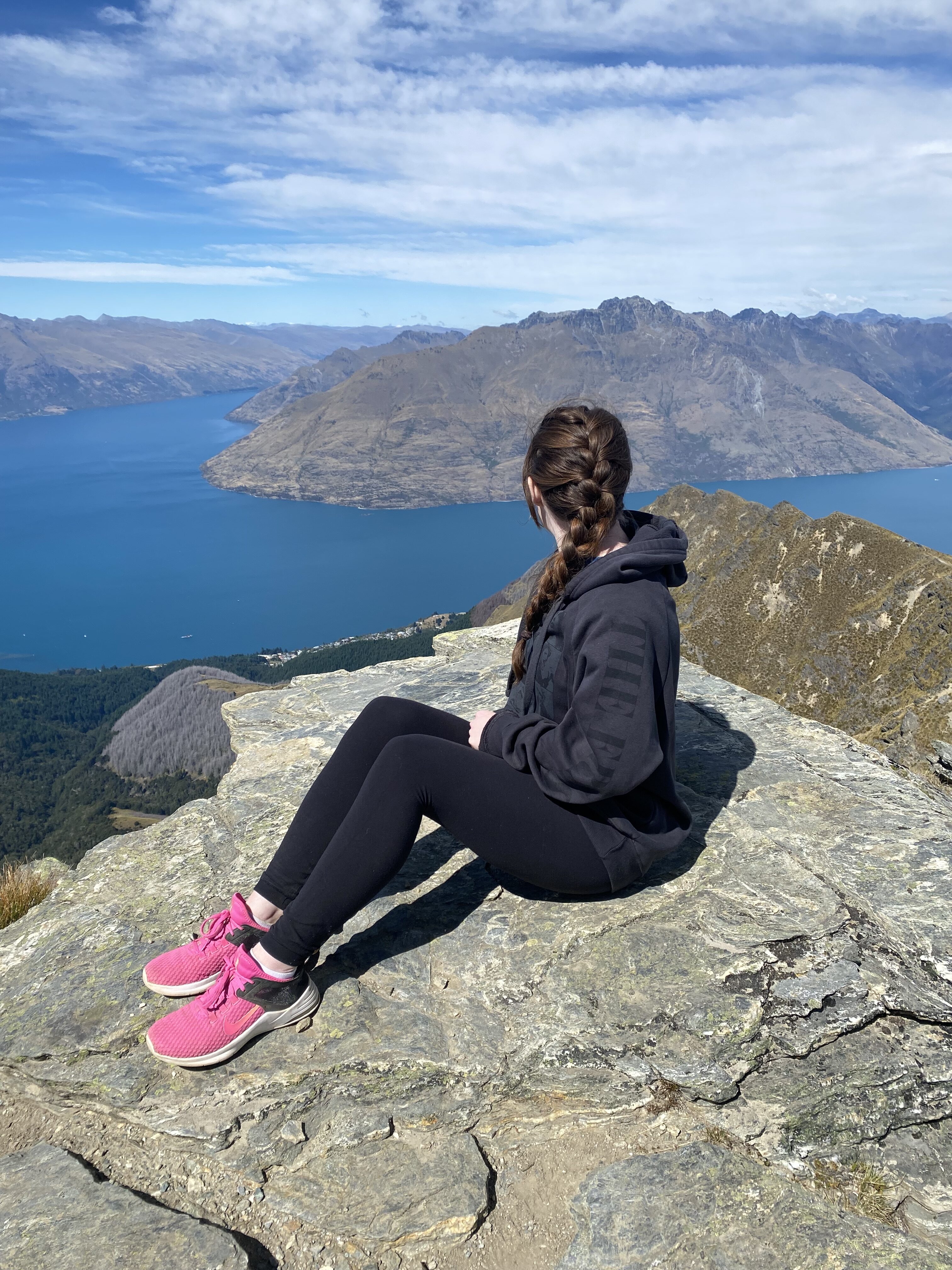 A hiker wearing pink shoes, black leggings and a dark grey sweatshirt looking out on Lake Wakatipu from the top of Queenstown.