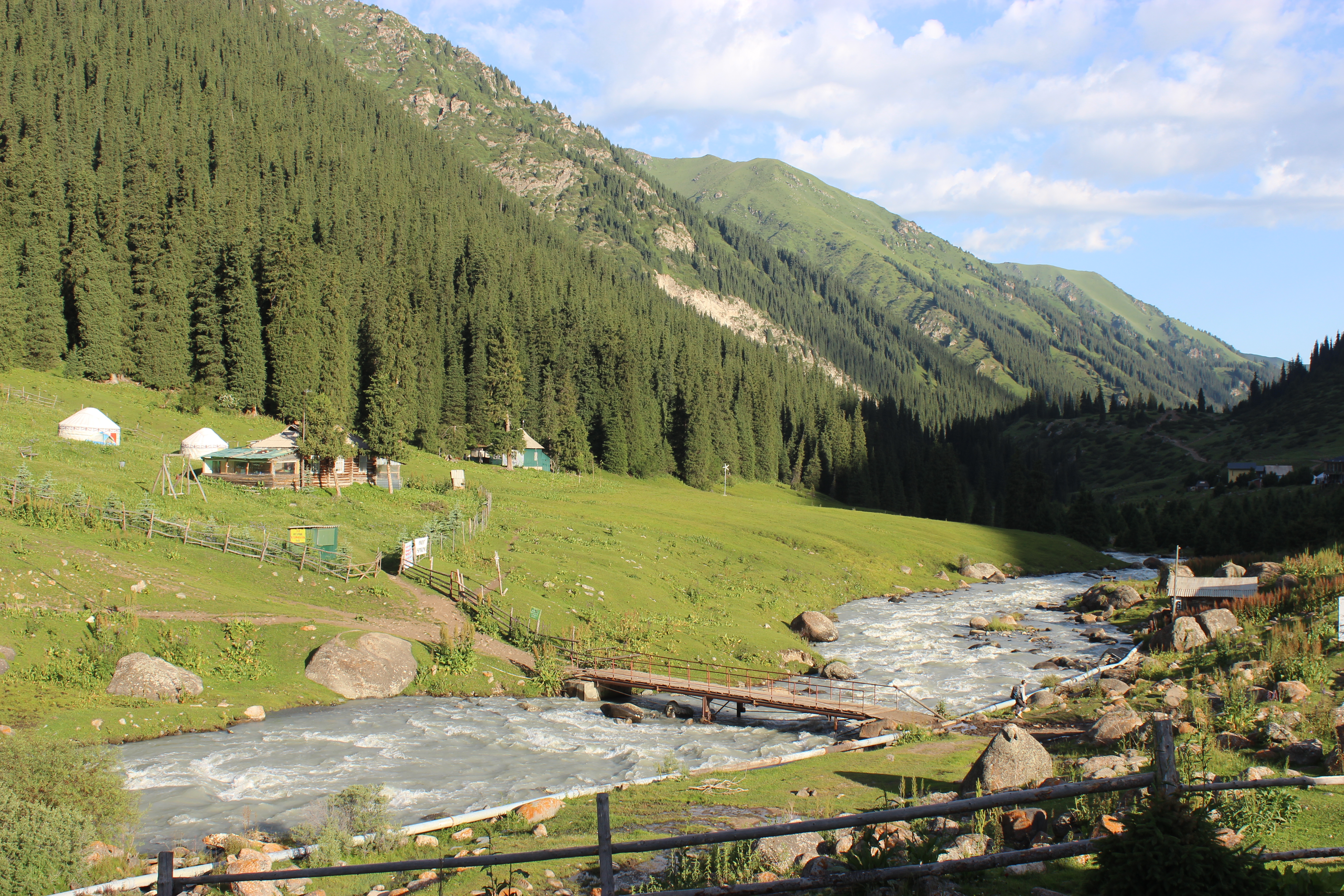 A river with a wooden bridge running through the Altyn Arashan valley with yurt camps on the hillside near the river banks.